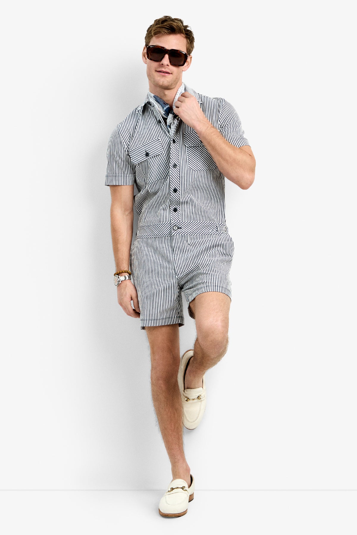 A man in the Sherman Short Jumpsuit, sunglasses, a watch, a blue patterned scarf, and cream loafers stands against a white background, leaning with one leg crossed.