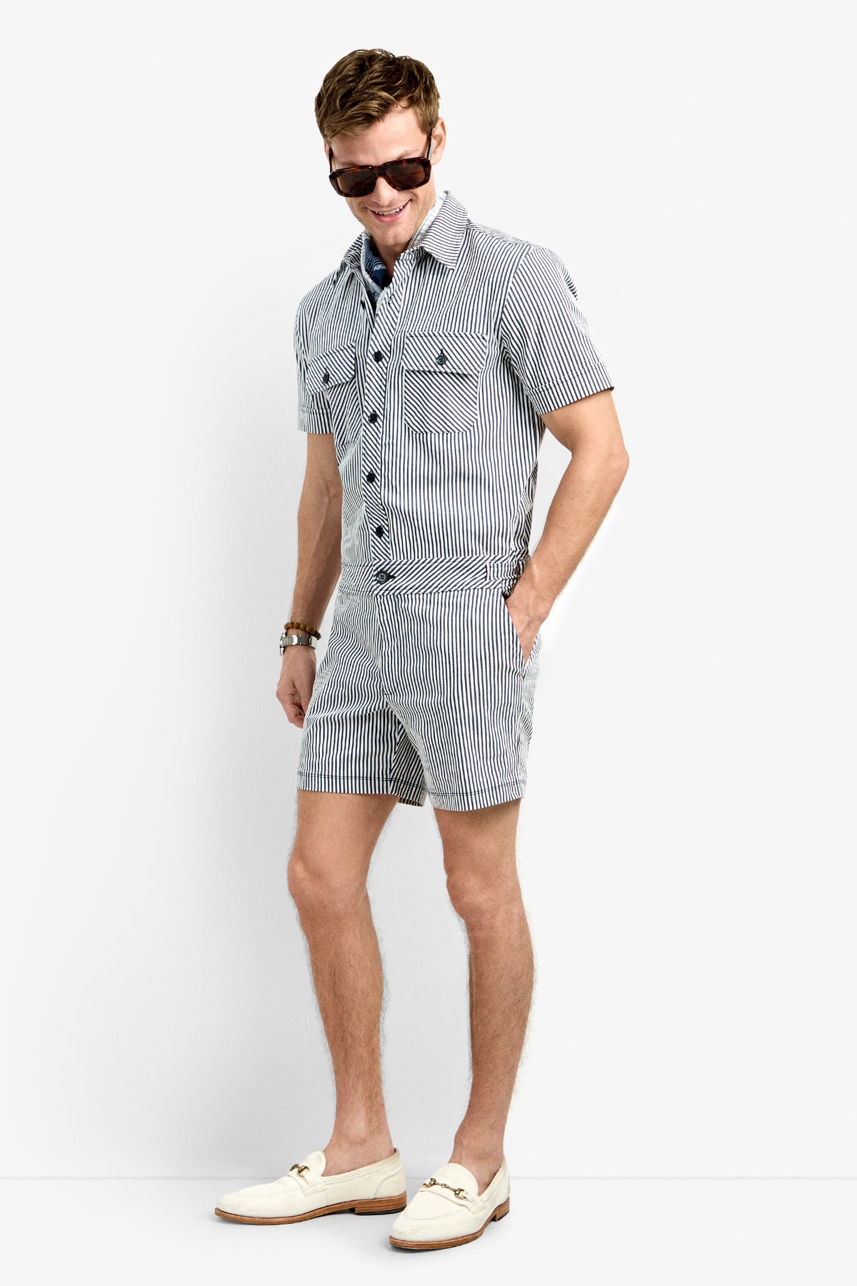 A man smiles with one hand in his pocket, wearing the Sherman Short Jumpsuit—a black-and-white striped, short-sleeve, button-up outfit—paired with sunglasses, white loafers, and a bracelet.