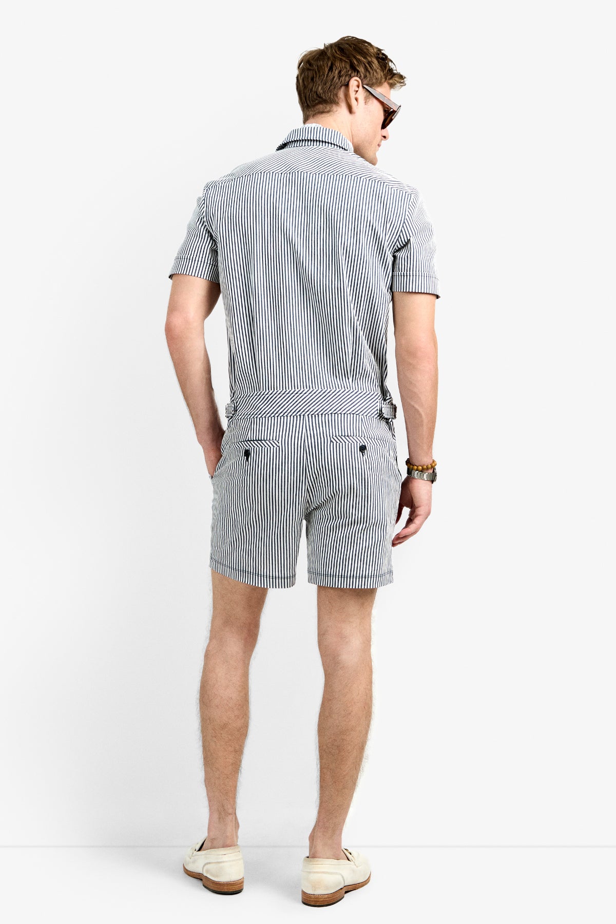 A man stands facing away, wearing the Sherman Short Jumpsuit—a black and white striped short-sleeve outfit—with sunglasses and light-colored shoes.