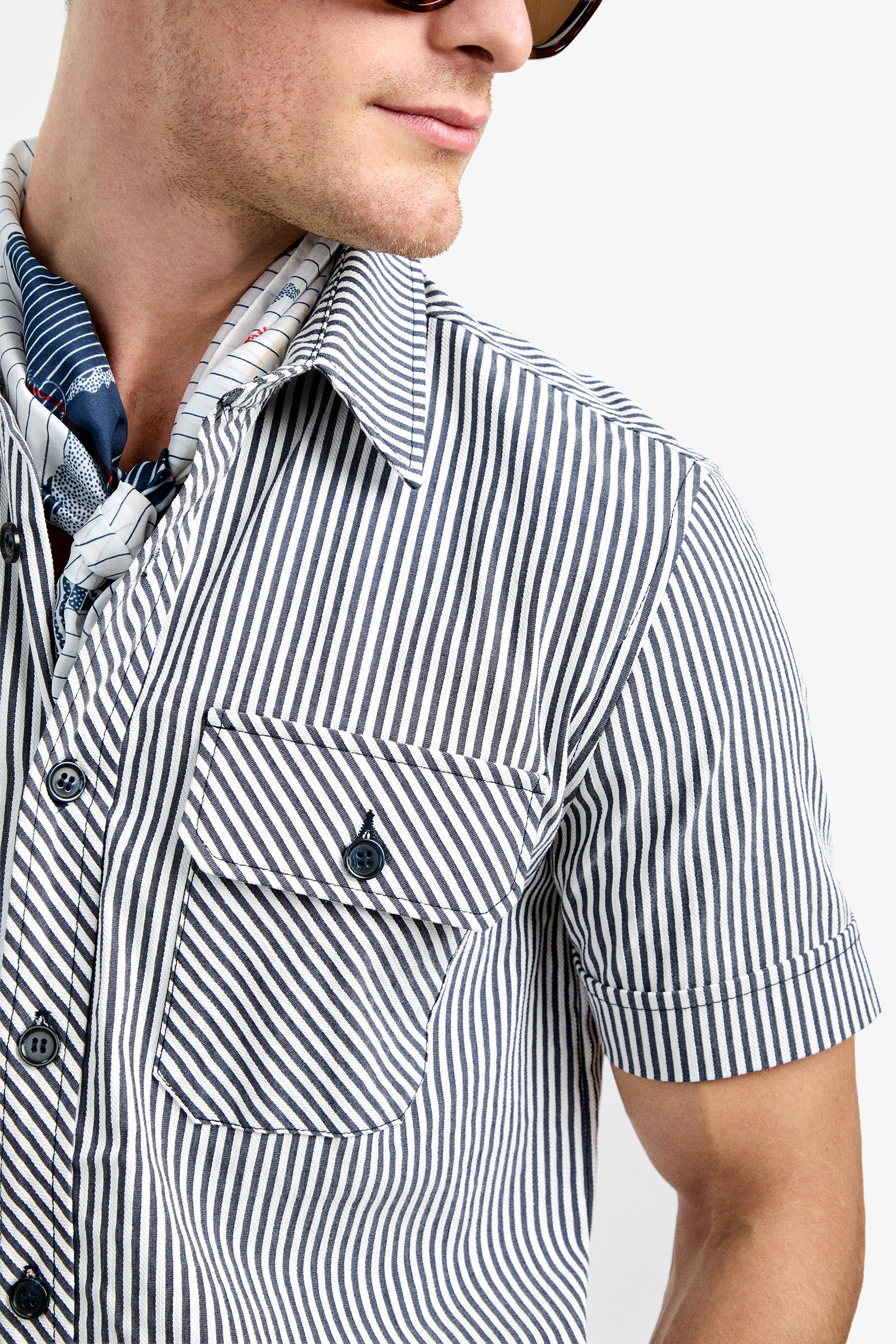 A person wearing sunglasses and the Sherman Short Jumpsuit with a blue and white striped pattern, short sleeves, front pocket, and a neck scarf, looks to the side against a plain background.