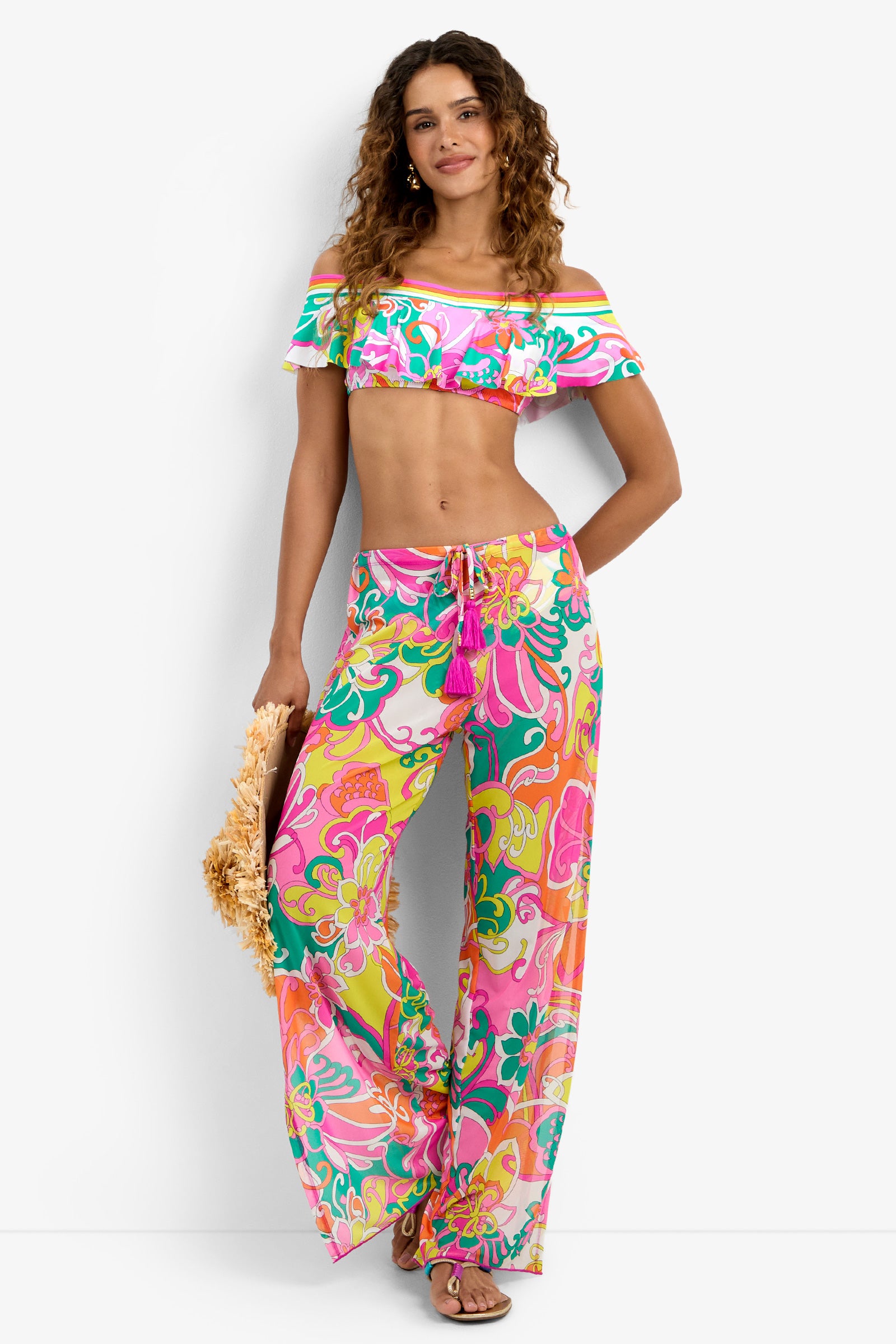 Woman wearing the Off The Shoulder Ruffle Bandeau Swim Top in MULTI, styled with wide-leg pants, a straw handbag, and sandals, smiling against a white background.