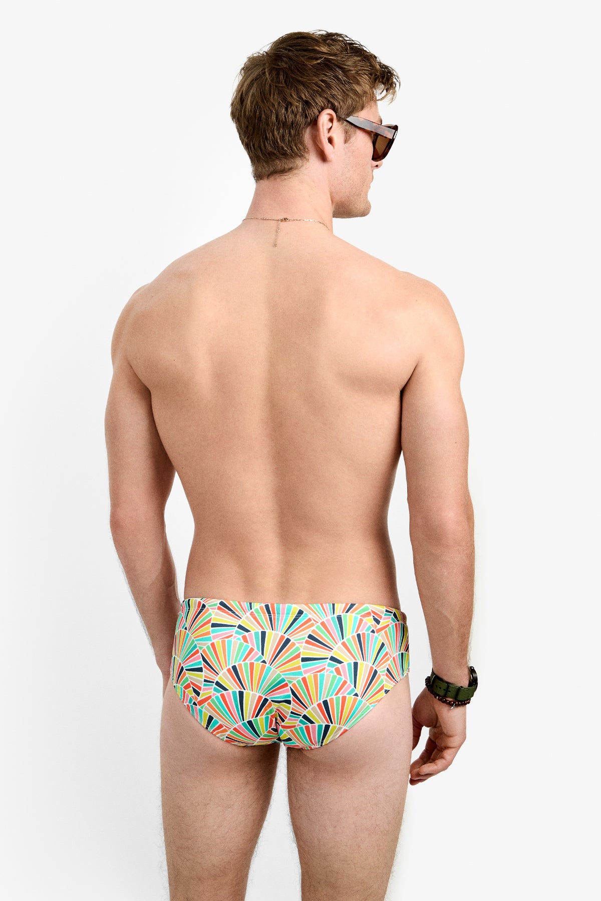 A man, seen from behind, models the Bondi Swim Brief with a colorful geometric pattern, sunglasses, and a wristwatch against a plain white background.