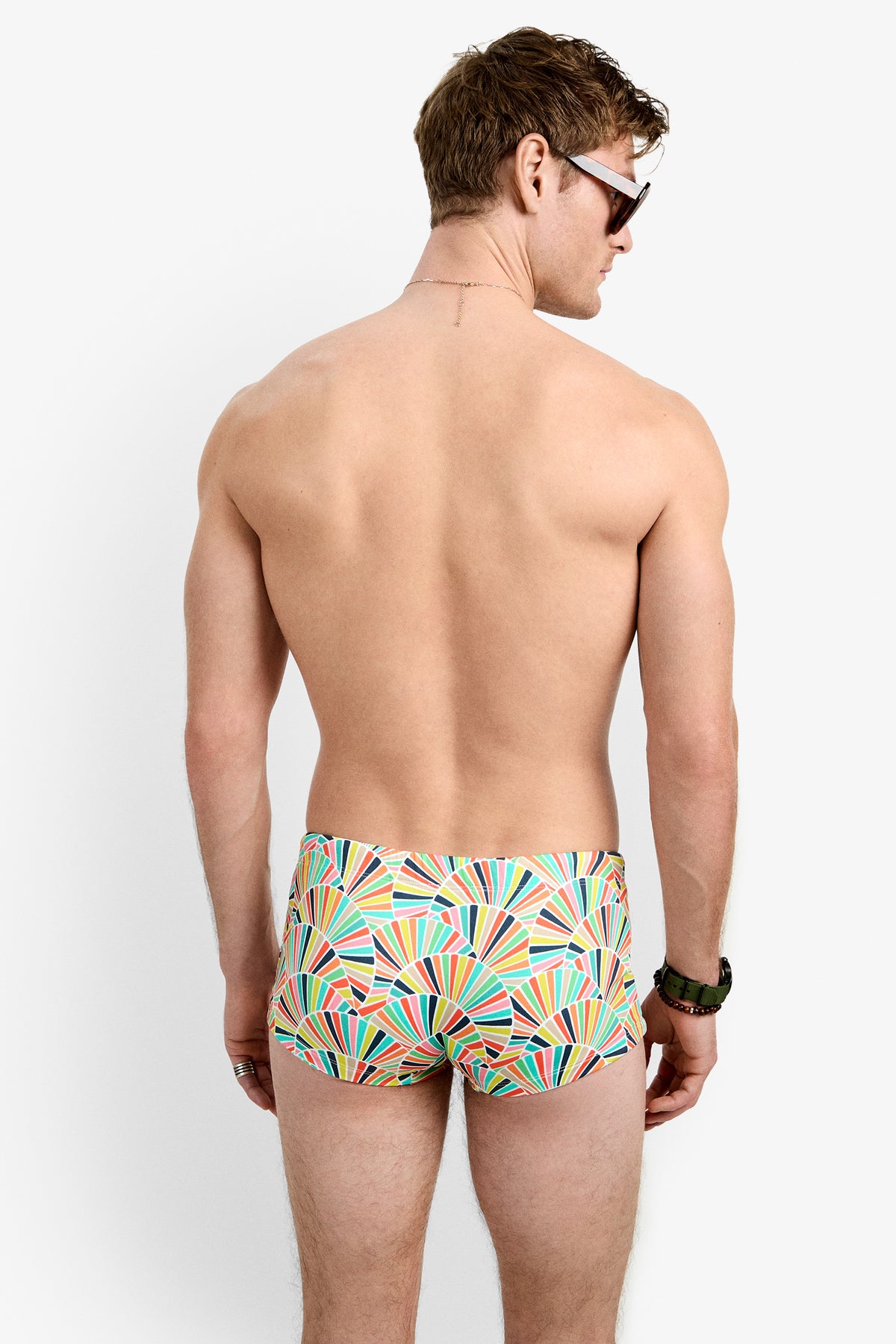A man stands with his back to the camera, wearing Sorrento Swim Trunk featuring colorful geometric patterns, along with sunglasses and a watch, set against a plain white background.