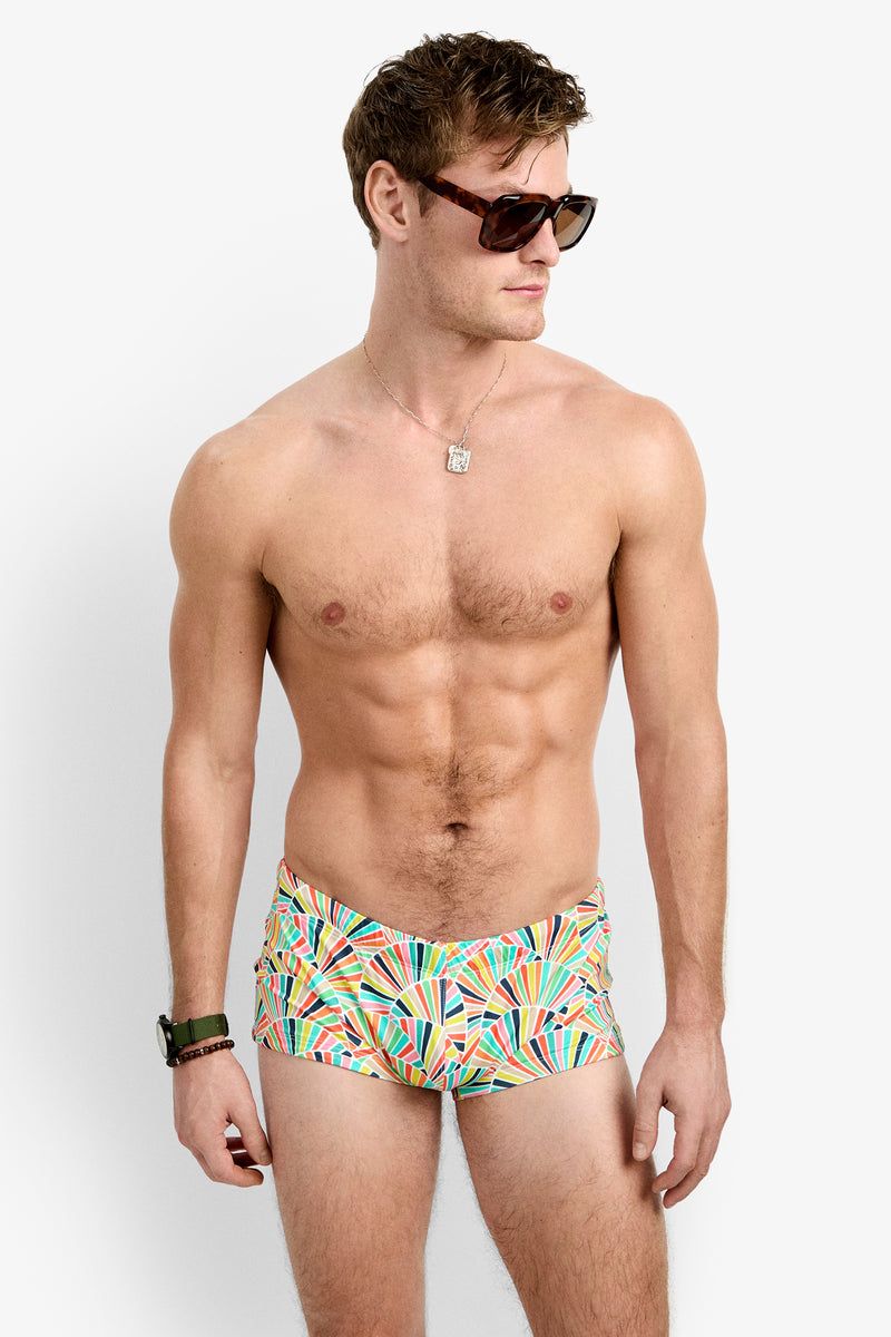 A man wearing Sorrento Swim Trunk with colorful geometric patterns, sunglasses, a wristwatch, and a necklace stands against a plain white background, looking to his right.