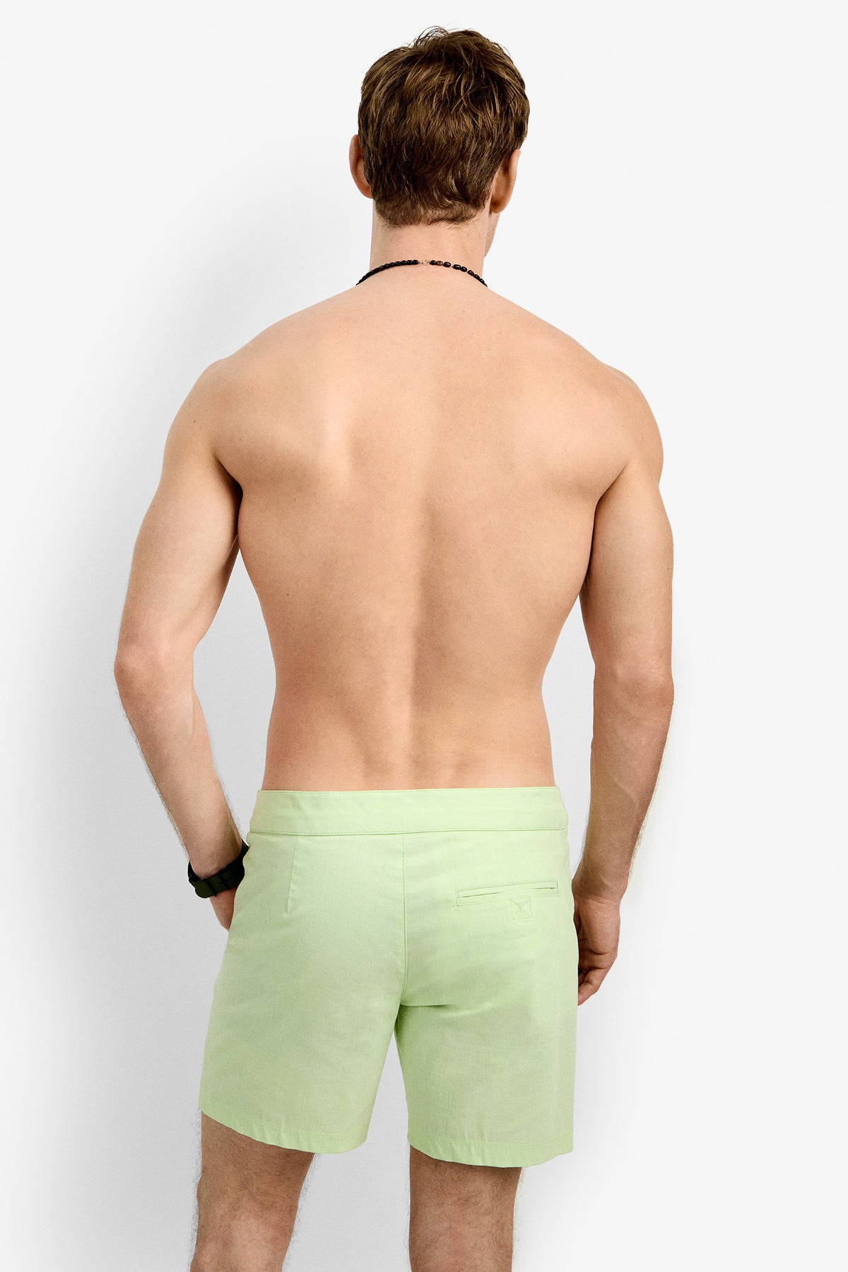 A shirtless, light-skinned man with brown hair stands facing away in Honolulu Board Short (light green) and a black wristband, set against a plain white background.