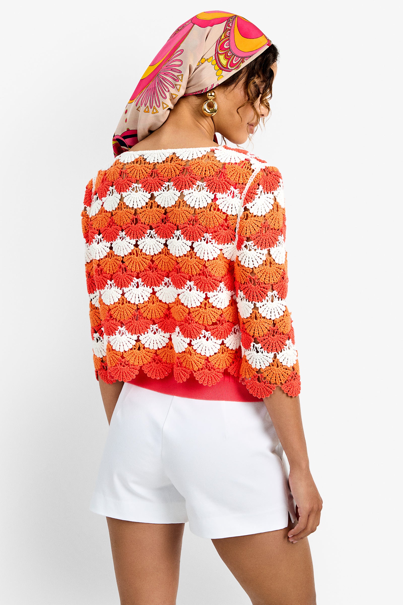 A woman with a colorful headscarf and hoop earrings models the Sofia Cardigan, a red, orange, and white crocheted top with scalloped designs, paired with white shorts as she stands with her back to the camera against a plain background.