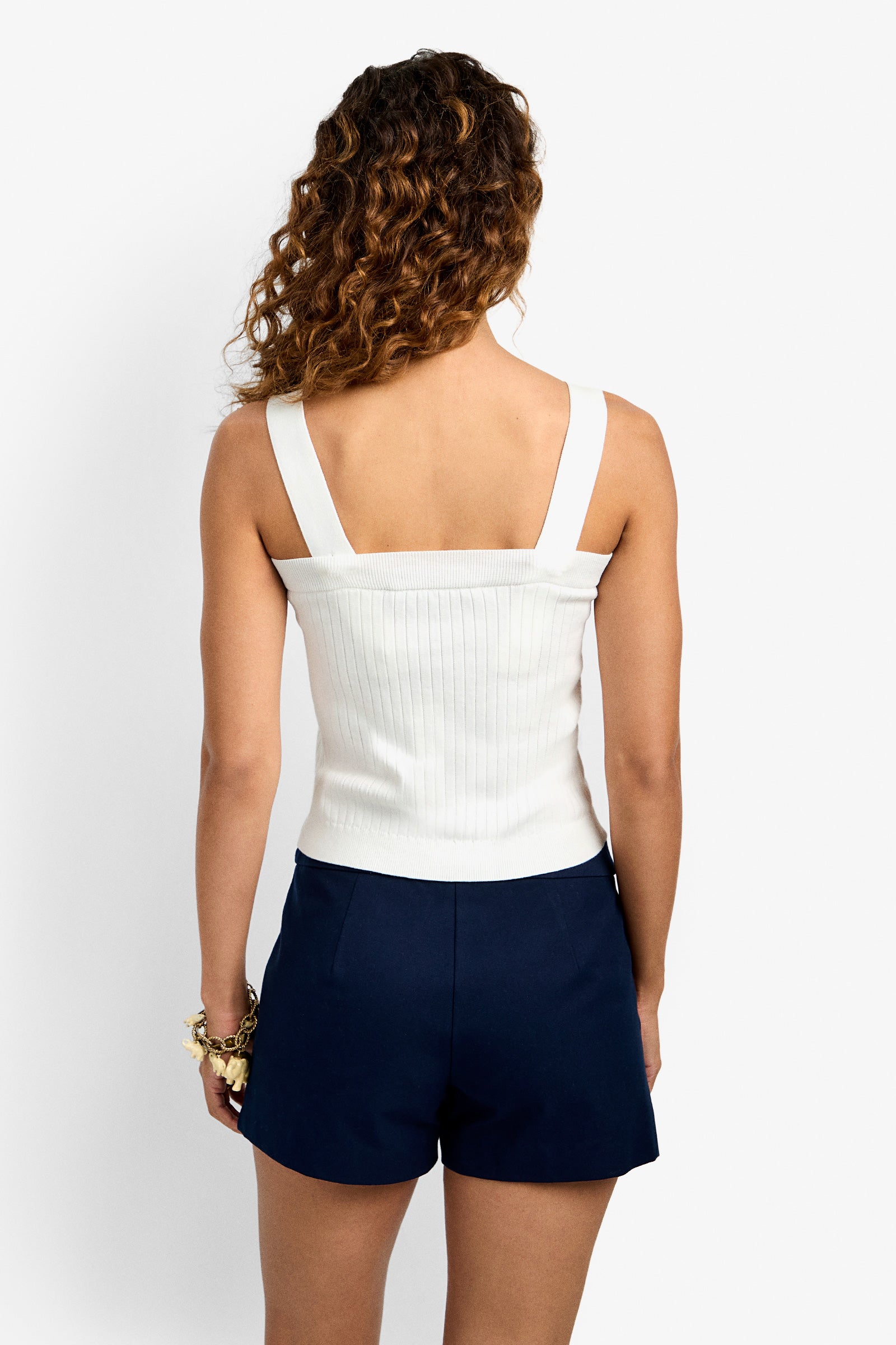 A woman with curly hair is seen from behind wearing the Breakpoint Cami, a white ribbed sleeveless top, paired with navy blue shorts and a bracelet on her left wrist, standing against a plain white background.