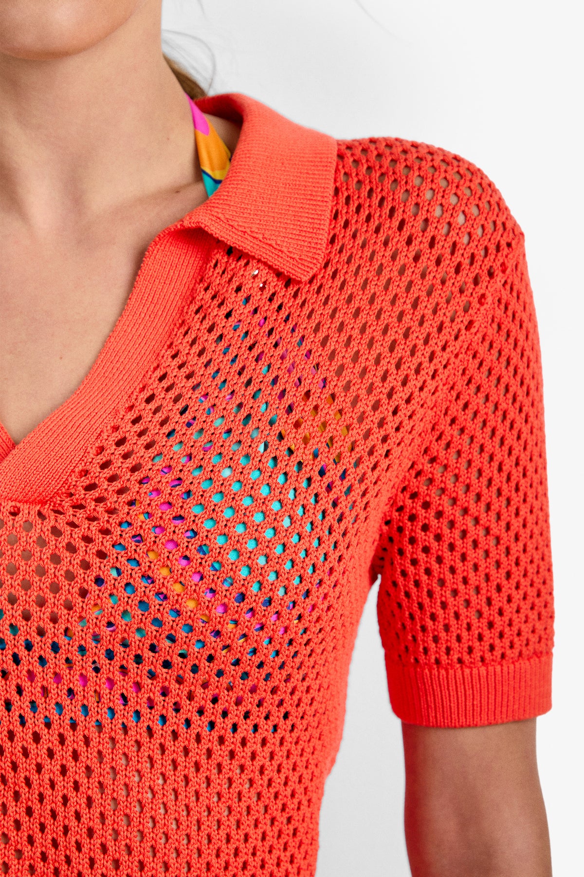 Someone is wearing the Jadelyn Mest Polo, a bright coral crochet-knit, short-sleeve shirt with a collar. A colorful swimsuit in blue, pink, and yellow is visible underneath through the mesh fabric. Only the upper torso is shown.