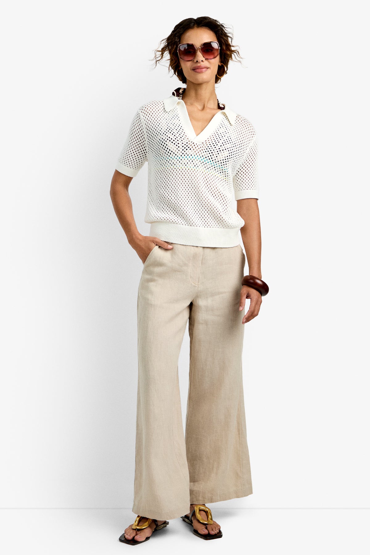 A woman wearing the Jadelyn Mest Polo, white camisole, beige wide-leg pants, brown sandal flats, and a large brown bracelet stands with one hand in her pocket and smiles.
