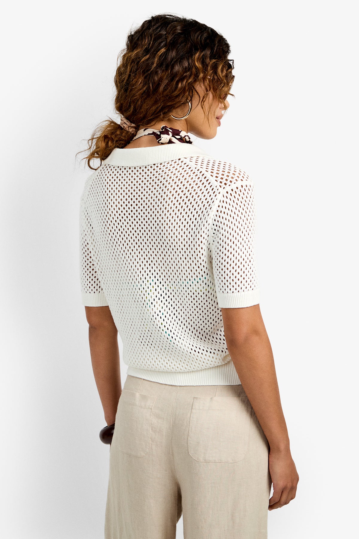 A woman with a loose ponytail faces away from the camera, wearing the Jadelyn Mest Polo—a white short-sleeve mesh sweater—over a bikini top and light beige pants. The setting features a plain white background.