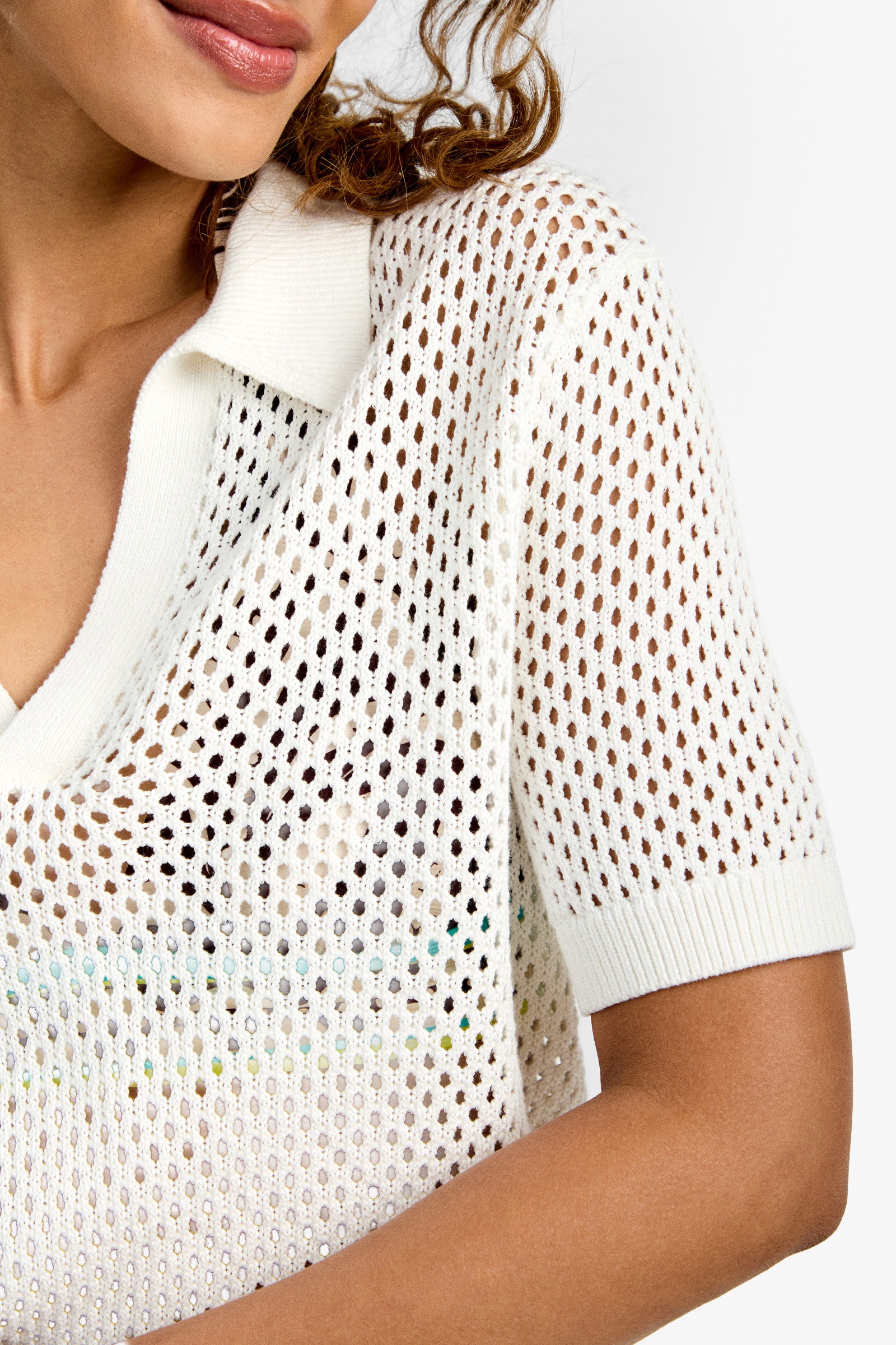 A woman softly smiles in the Jadelyn Mest Polo, a white short-sleeve, collared open-knit top with a deep V-neck, her curly hair partly visible.