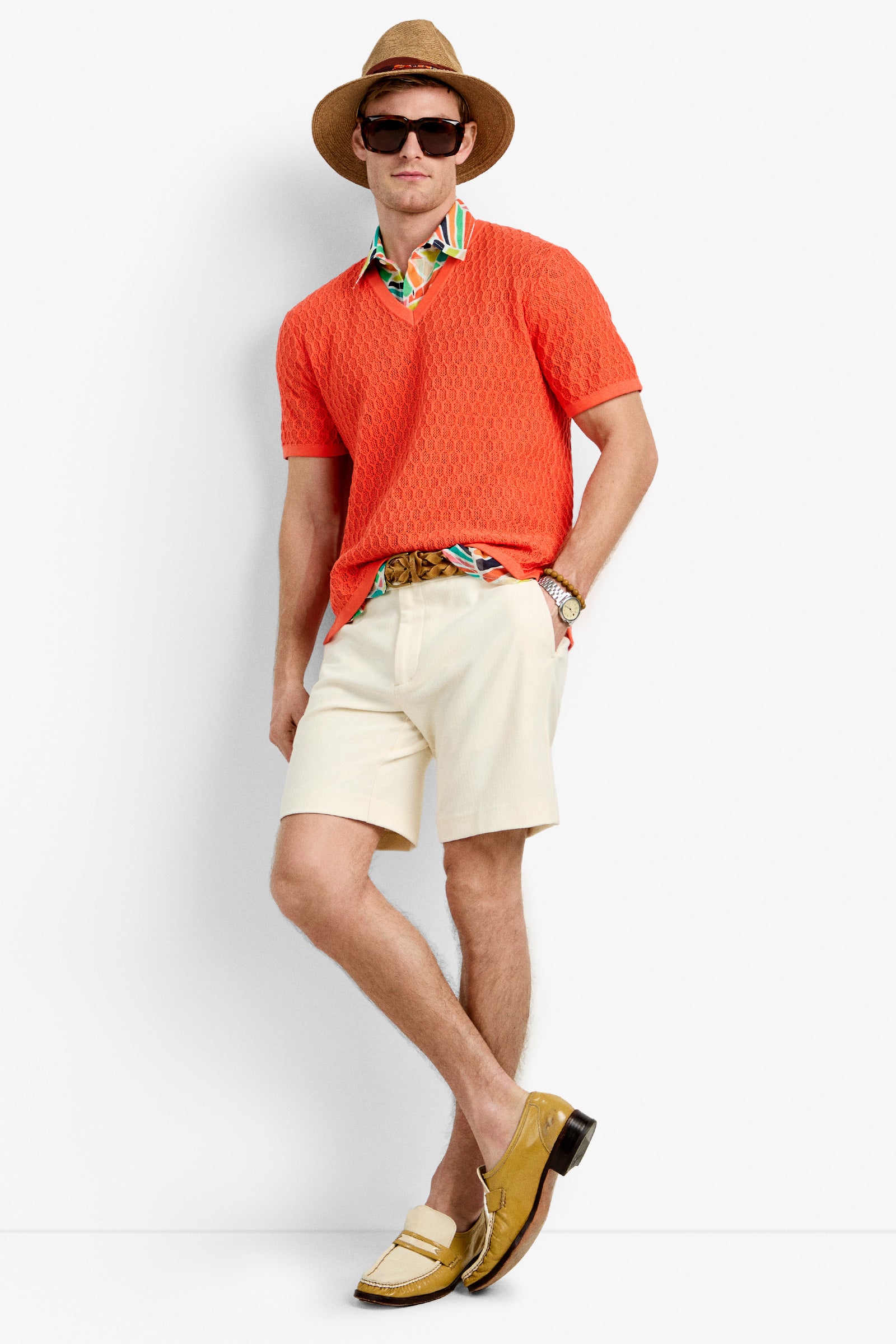 A man wearing the Heyes Mesh Henley V-Neck, a brown hat, dark sunglasses, cream shorts, a brown woven belt, a watch, and yellow loafers stands against a white background.