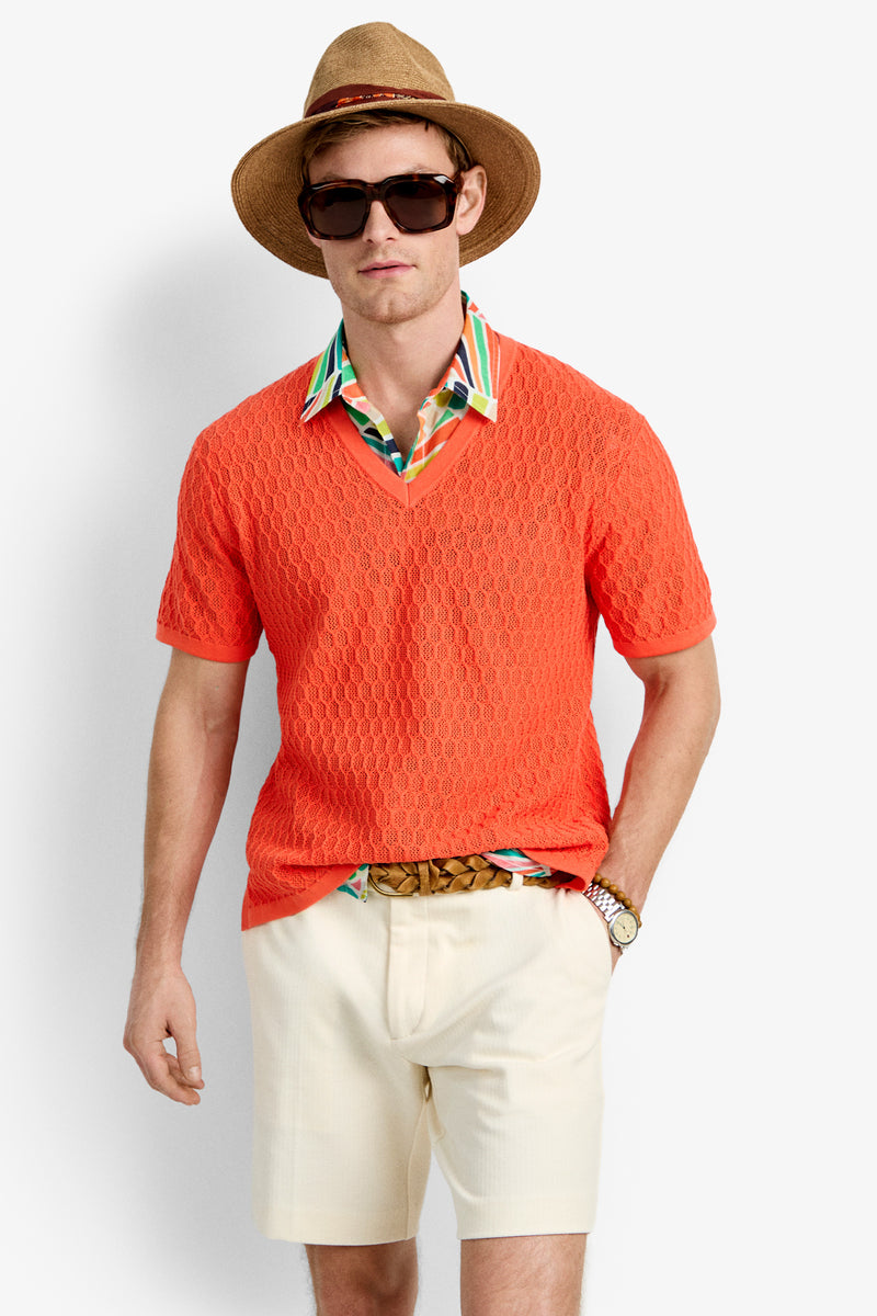 A man wearing the Heyes Mesh Henley V-Neck over a colorful collared shirt, paired with cream shorts, a brown woven belt, dark sunglasses, a straw hat, and a wristwatch stands against a plain white background.