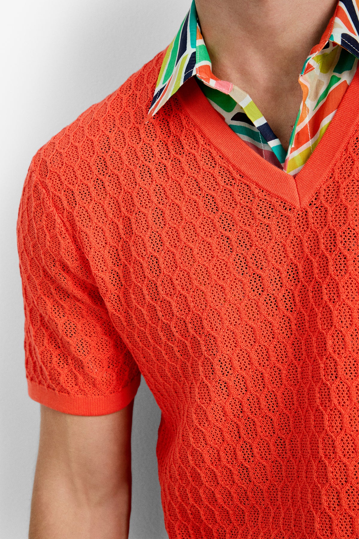A person is wearing the Heyes Mesh Henley V-Neck, a bright orange short-sleeve knit sweater layered over a colorful patterned collared shirt with green, yellow, blue, and white shapes. Only their upper torso and part of their face are visible.