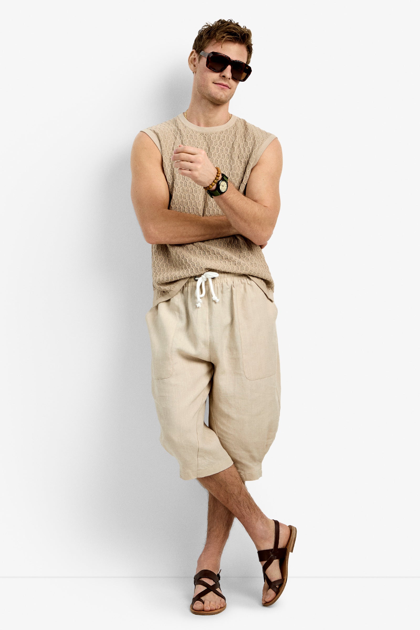 A man in Tovey Mesh Tan sunglasses, a beige knit sleeveless top, loose beige drawstring shorts, a wristwatch, and brown sandals stands against a white background giving a relaxed thumbs-up.