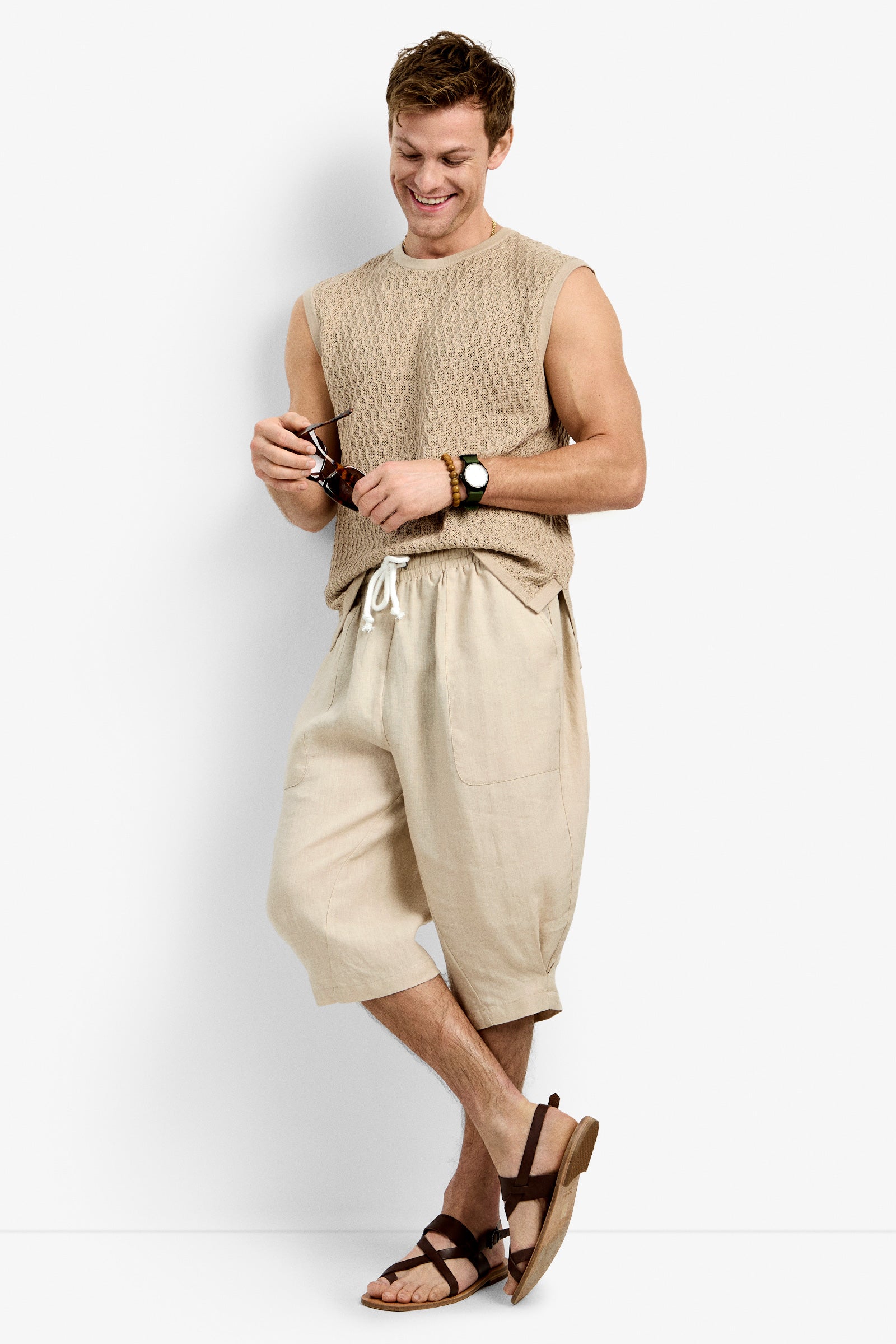 A man smiles, wearing a sleeveless beige knit top, loose light beige shorts, brown Tovey Mesh Tan sandals, a wristwatch, and holding sunglasses. He stands with relaxed arms and one leg crossed over the other.