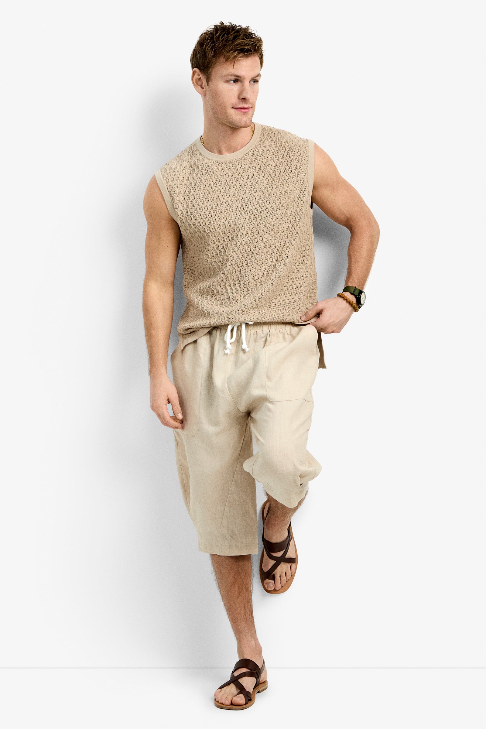 A man models the Tovey Mesh Tan in BEACHY BEIGE—a sleeveless textured top with loose, light drawstring pants—paired with brown sandals, standing against a white background with one leg crossed.