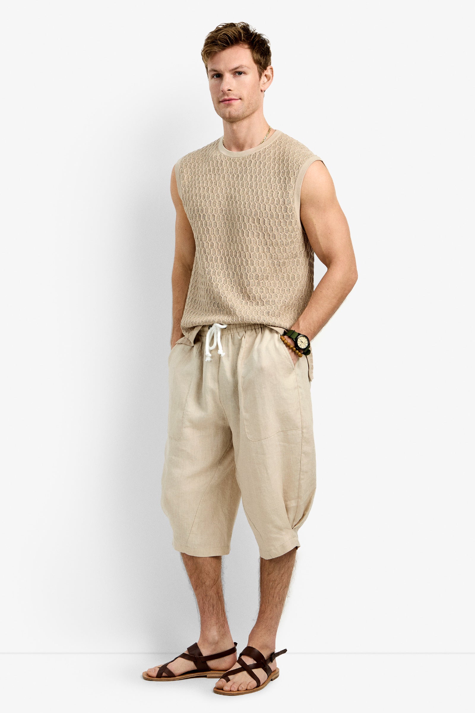 A man stands against a plain white background wearing Tovey Mesh Tan sleeveless knit top, loose light tan drawstring shorts, brown sandals, and a watch, with his hands in his pockets.