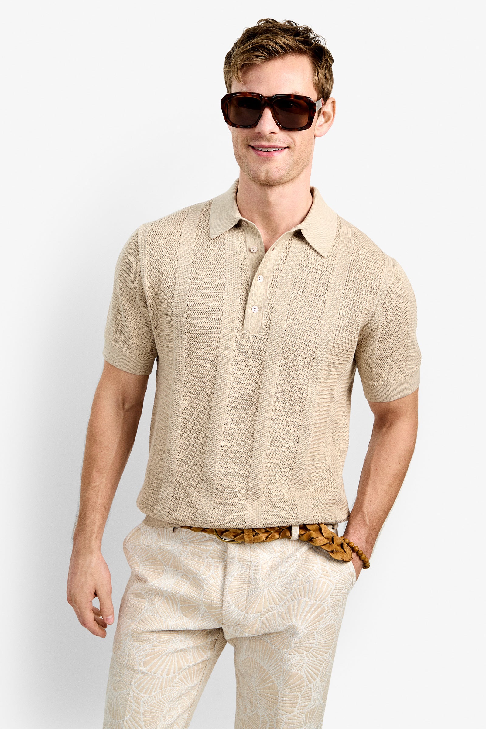 A man wearing oversized dark sunglasses, the Devan Short Sleeve Polo in beige, and light patterned trousers with a brown belt stands smiling with one hand in his pocket against a plain light background.