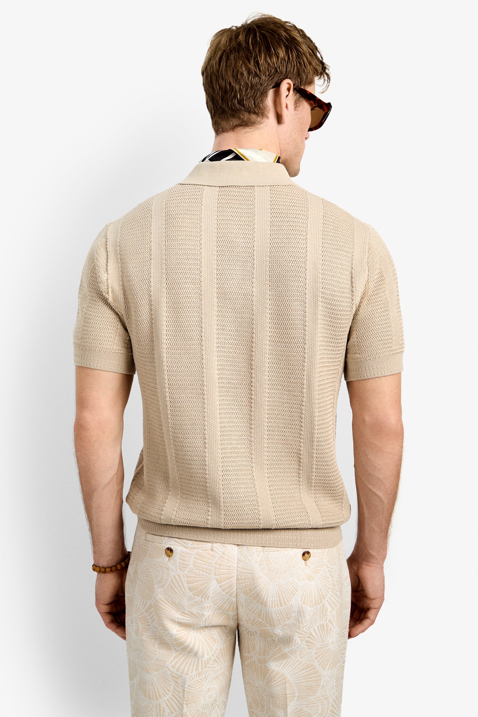 A man with short light brown hair is seen from behind wearing the Devan Short Sleeve Polo in beige, paired with light patterned trousers, sunglasses, and a scarf tucked into his collar.