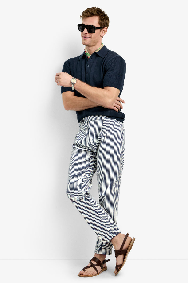 A man wearing the Devan Short Sleeve Polo stands against a white background with sunglasses, striped pants, brown sandals, arms crossed, and legs casually crossed.