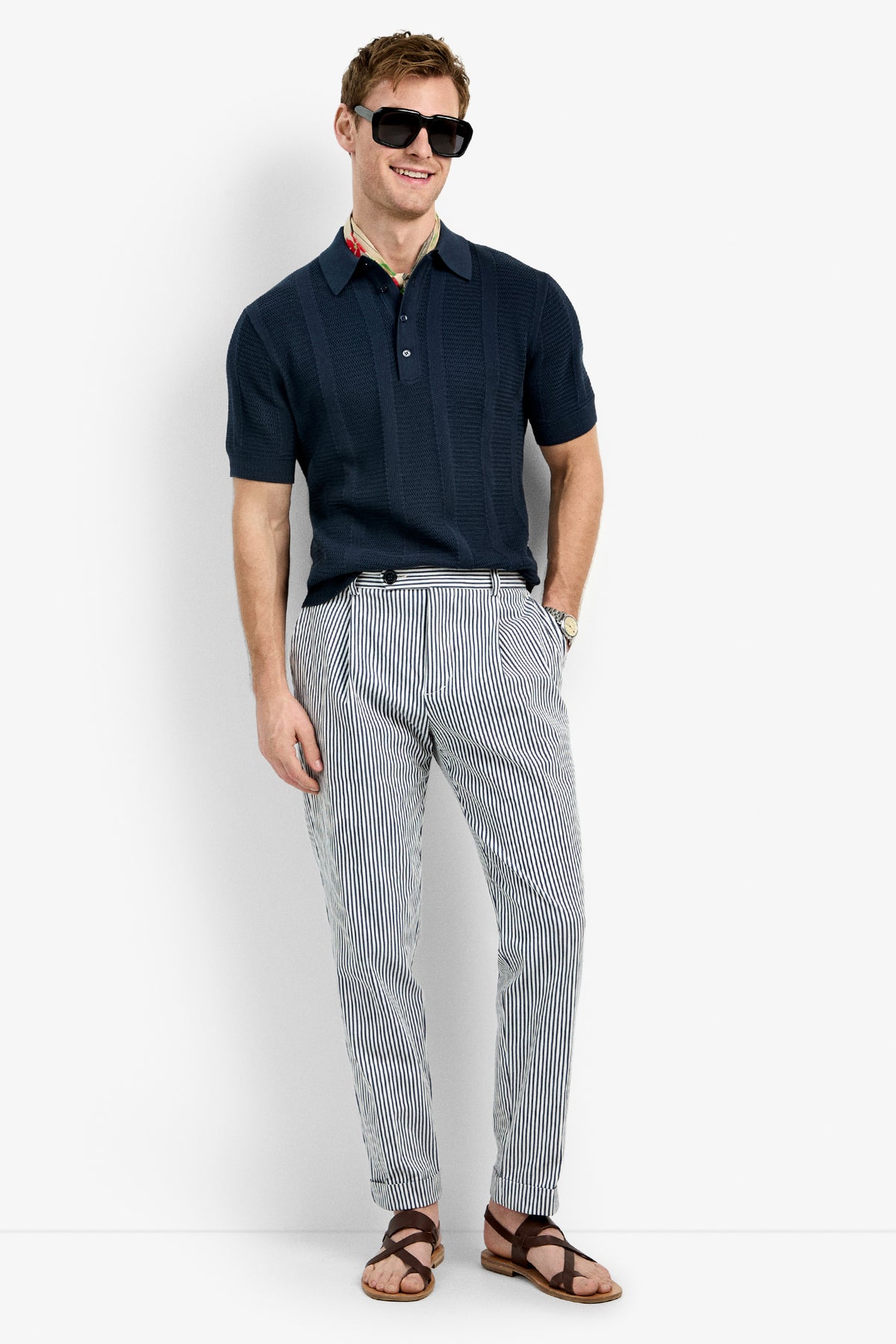 A man smiles with one hand in his pocket, wearing the Devan Short Sleeve Polo in navy, dark sunglasses, light striped pants, and brown sandals against a plain white background.