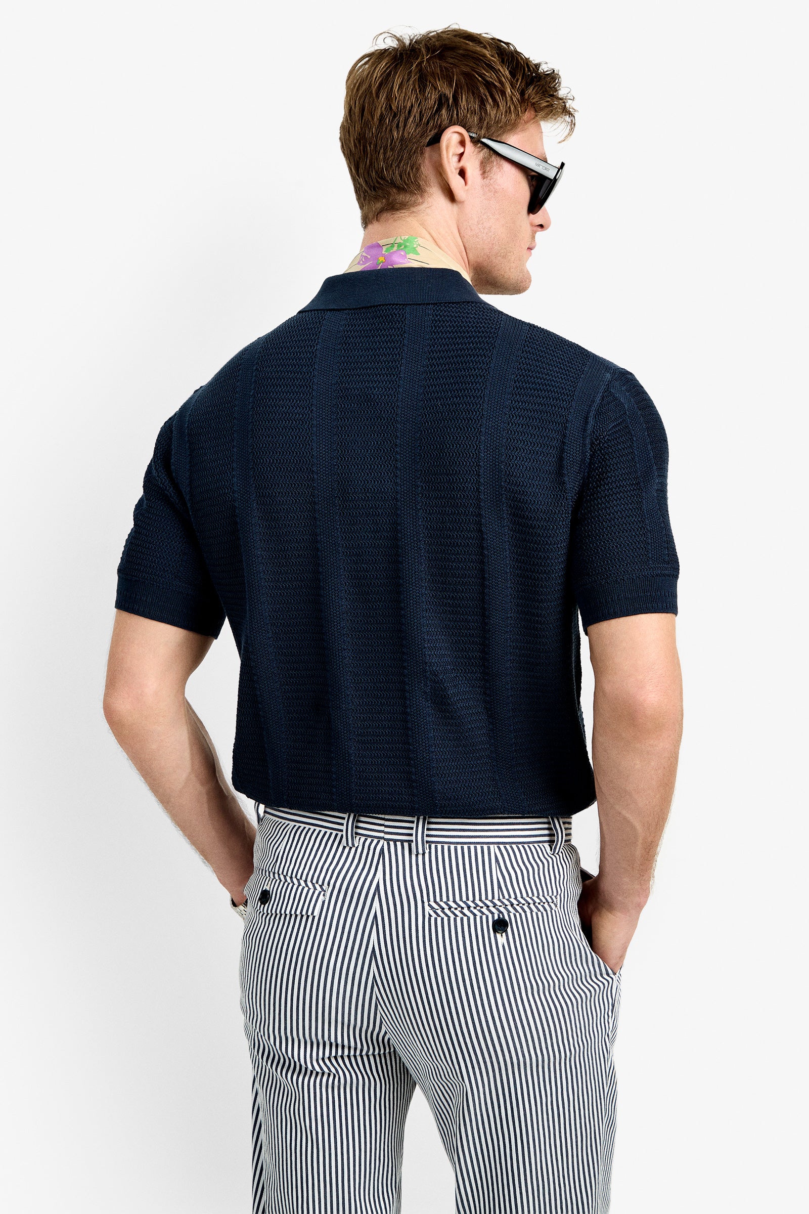 A man with short brown hair and sunglasses is seen from the back, wearing the Devan Short Sleeve Polo and high-waisted, black-and-white striped pants. He has a colorful neck tattoo and stands against a plain white background.