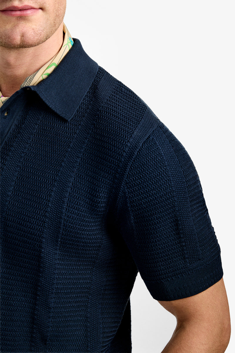 A man wears the Devan Short Sleeve Polo, a textured navy knit shirt layered over a patterned beige and green collared shirt, shown from mid-face to upper arm against a plain white background.