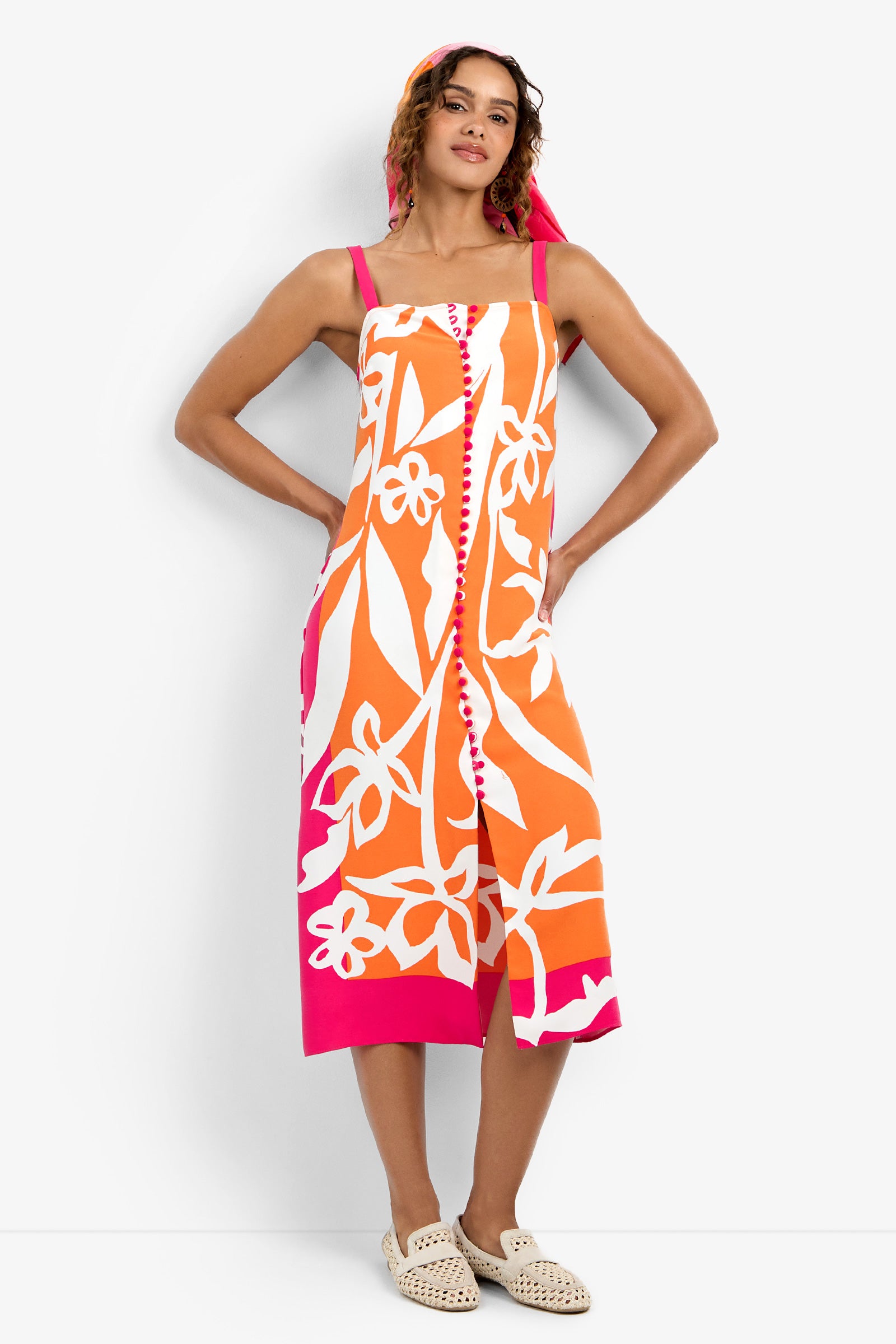 A woman poses with one hand on her hip, wearing the sleeveless Haisley Dress in orange and pink with bold white floral patterns, paired with a matching headscarf and cream woven slip-on shoes.