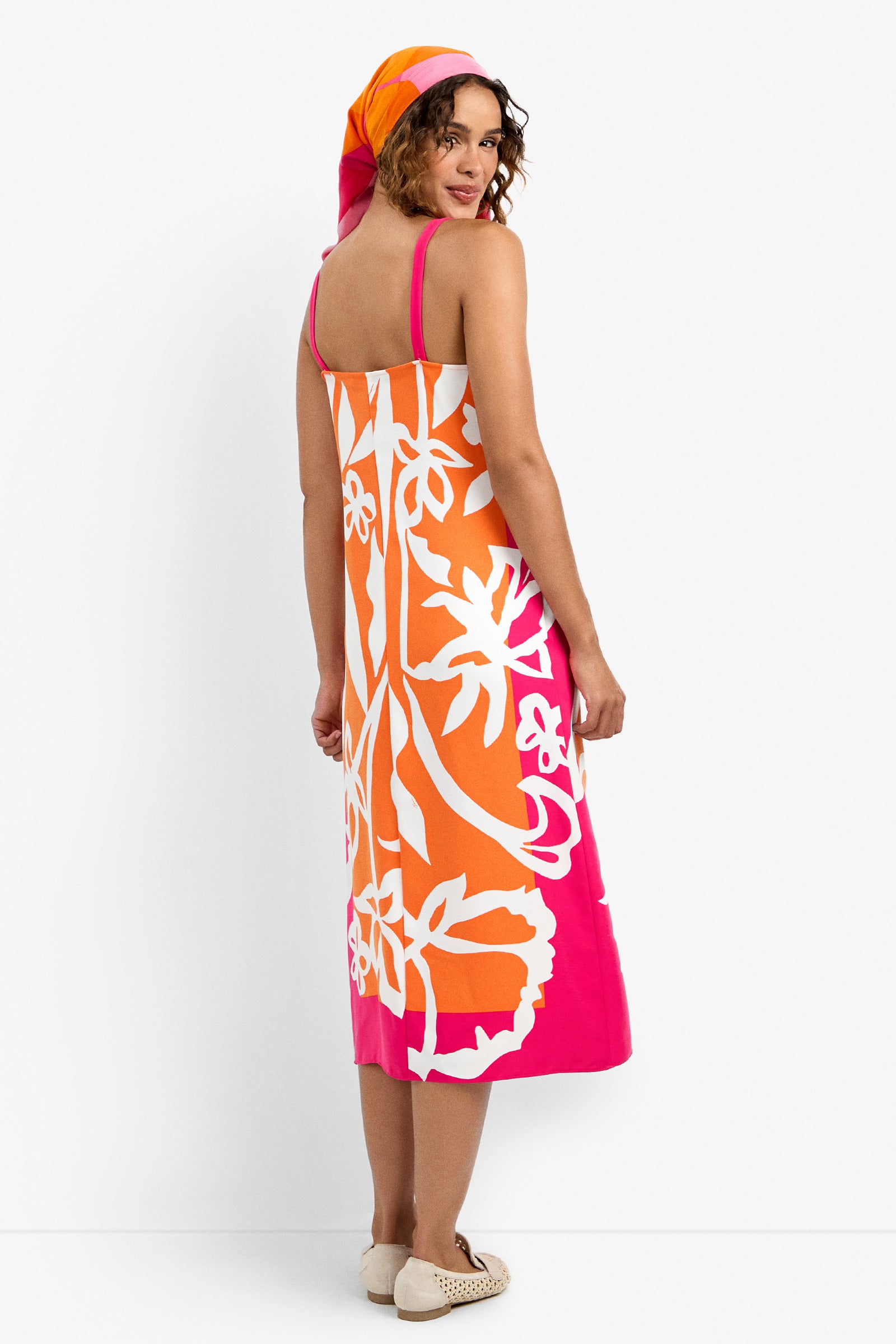 A woman, seen from behind, glances over her shoulder while wearing the sleeveless orange, pink, and white floral Haisley Dress with a matching headscarf and white flats against a plain white background.