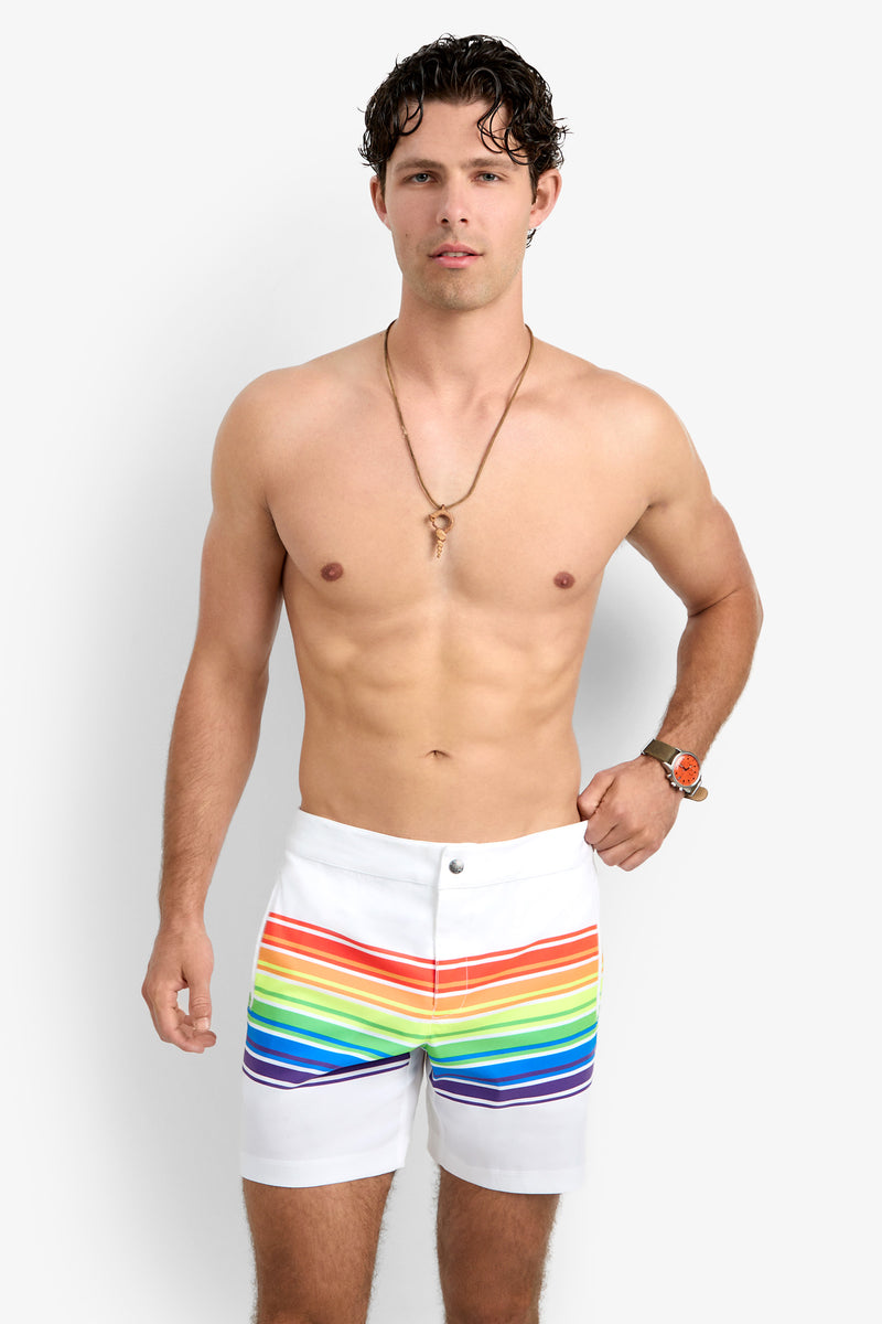 A shirtless man with short, wavy brown hair, a necklace, and an orange watch confidently models the Honolulu Board Short—white shorts with horizontal rainbow stripes—against a plain background.