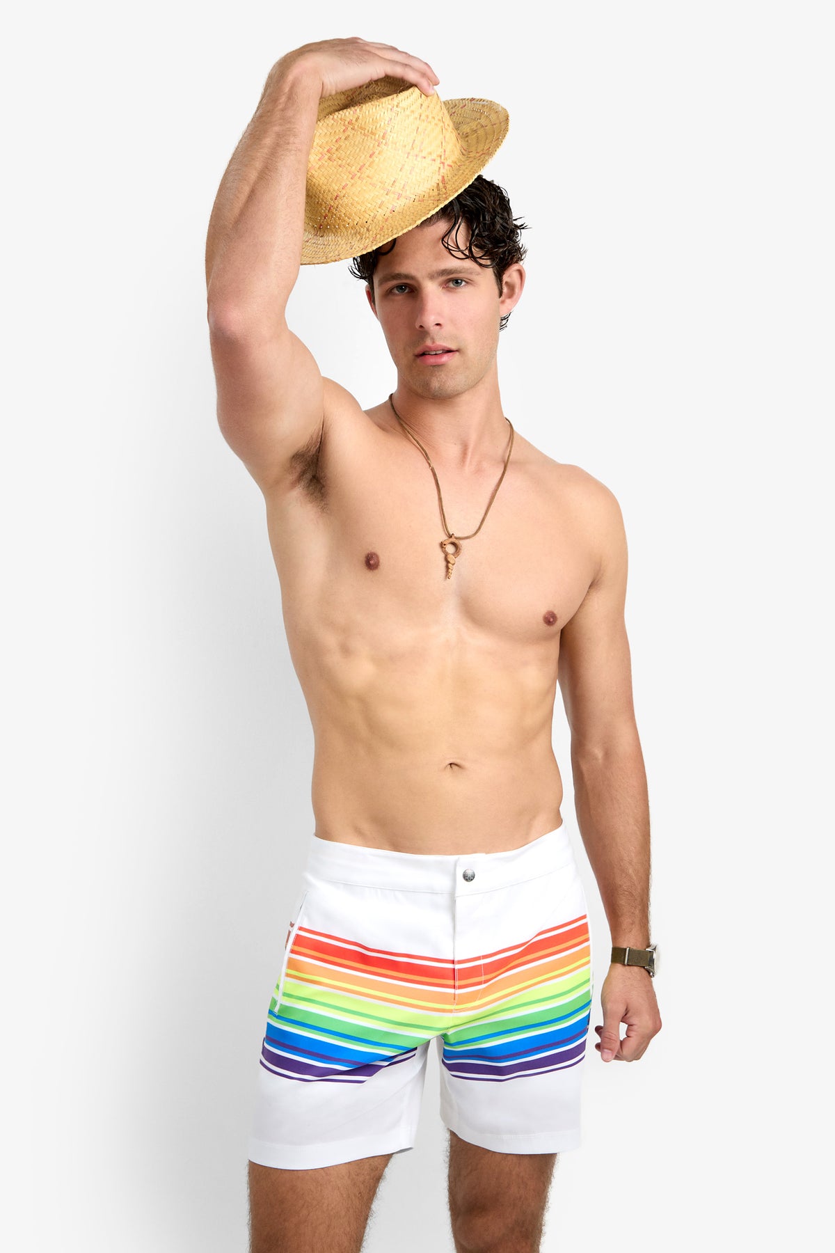 A man with short dark hair wears the Honolulu Board Short—white swim trunks with a rainbow stripe. He holds a straw hat above his head, accessorized with a necklace and wristwatch, standing against a plain white background.