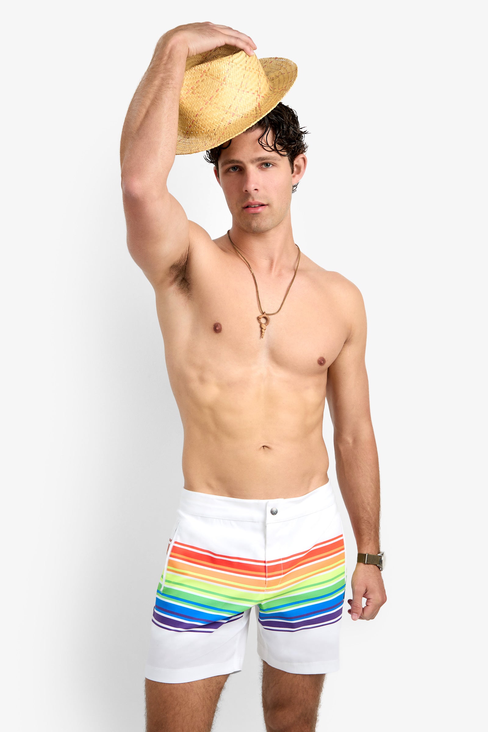A man with short dark hair wears the Honolulu Board Short—white swim trunks with a rainbow stripe. He holds a straw hat above his head, accessorized with a necklace and wristwatch, standing against a plain white background.