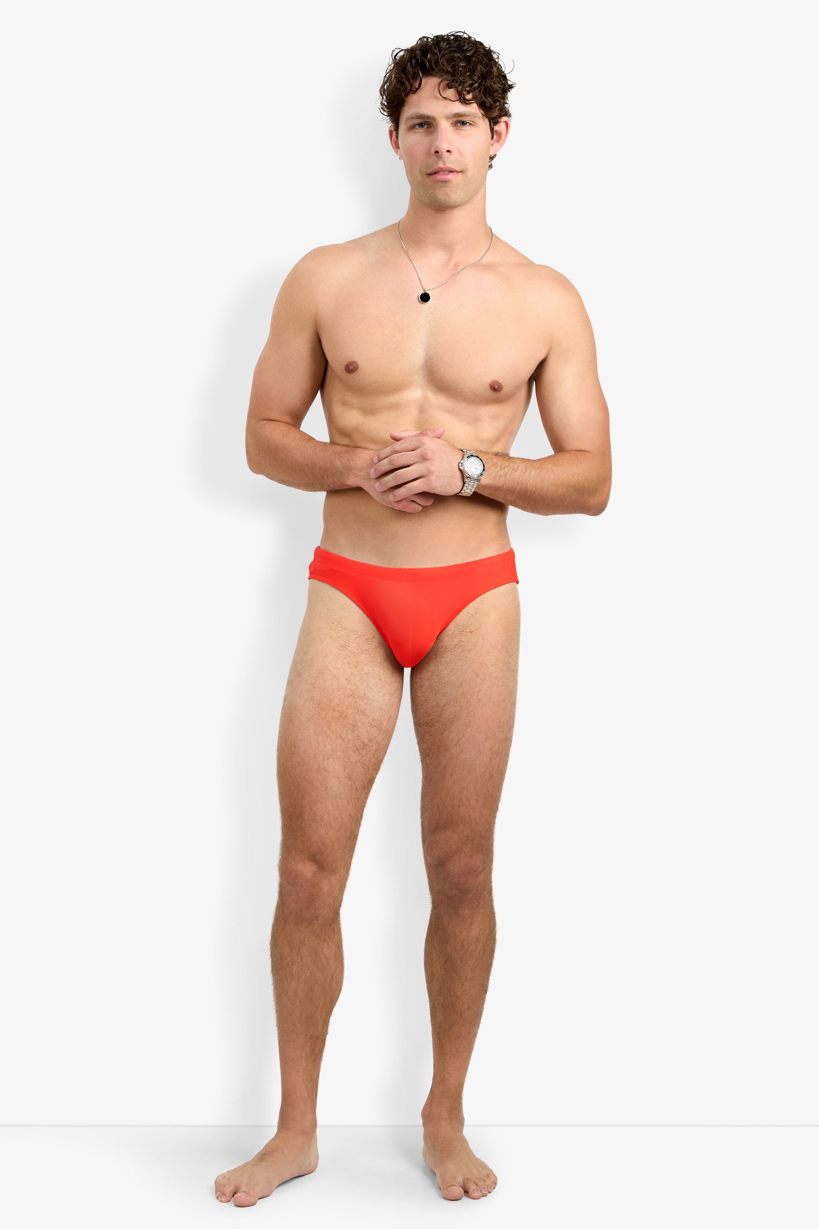 A man with curly hair stands barefoot against a white background, wearing the Bondi Swim Brief in bright orange, along with a wristwatch and necklace. His hands are clasped in front of him as he looks directly at the camera.