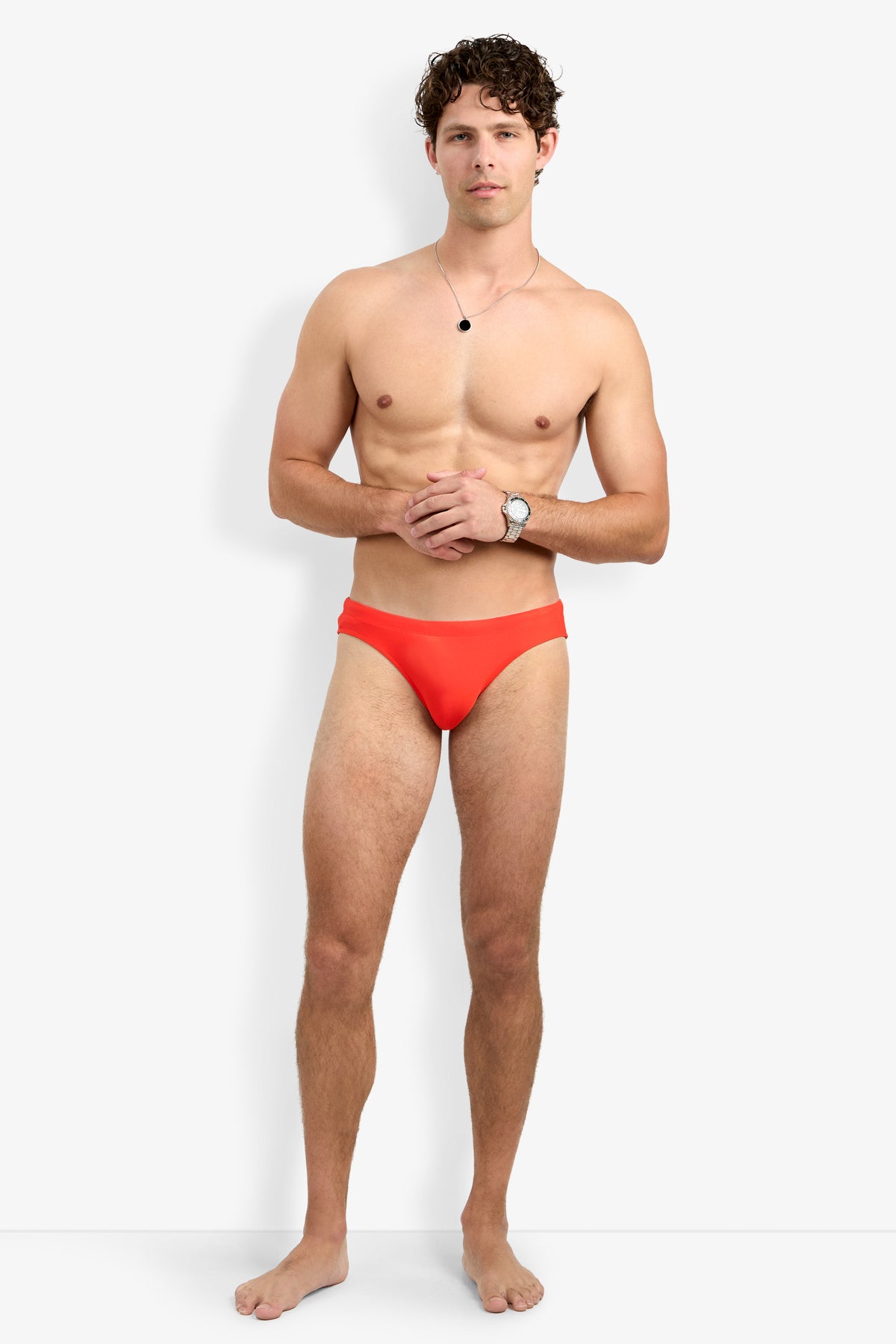 A young man with curly hair stands barefoot against a plain white background, wearing the Bondi Beach Swim Brief in bright orange, along with a wristwatch and necklace. He is shirtless, with his hands loosely clasped in front of him.