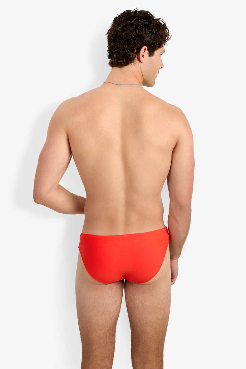 A man with short curly brown hair poses shirtless, facing away from the camera, wearing Bondi Beach Swim Briefs in bright red against a plain white background.