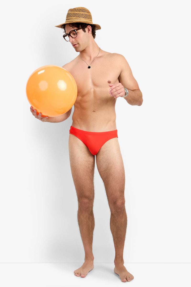 A man in the Bondi Beach Swim Brief, with a straw hat, glasses, and necklace, stands barefoot holding and pointing at an orange beach ball against a plain white background.
