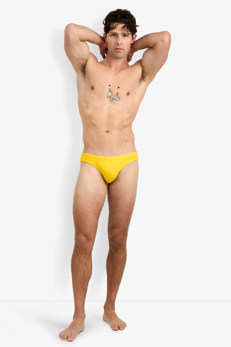 A man models the Bondi Swim Brief in yellow and a necklace, standing barefoot against a plain white background with his hands behind his head.