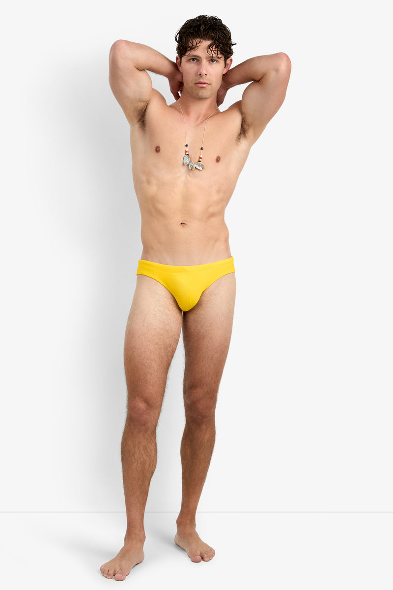 A man with curly hair stands barefoot against a white background, wearing the Bondi Beach Swim Brief in yellow and a necklace. His arms are raised, with his hands resting behind his head.