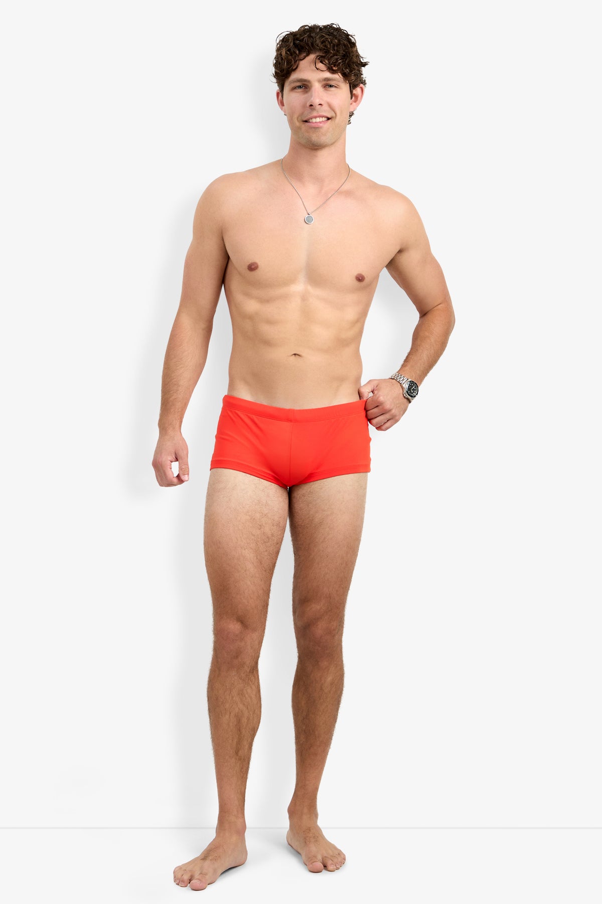 A smiling man with curly brown hair, barefoot and wearing a silver necklace, wristwatch, and Montego Bay Swim Trunk in bright orange stands against a plain white background with one hand relaxed by his side.