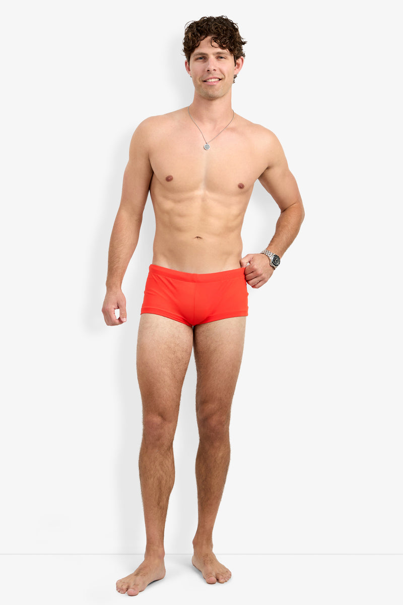 A smiling man with curly brown hair, barefoot and wearing a silver necklace, wristwatch, and Montego Bay Swim Trunk in bright orange stands against a plain white background with one hand relaxed by his side.