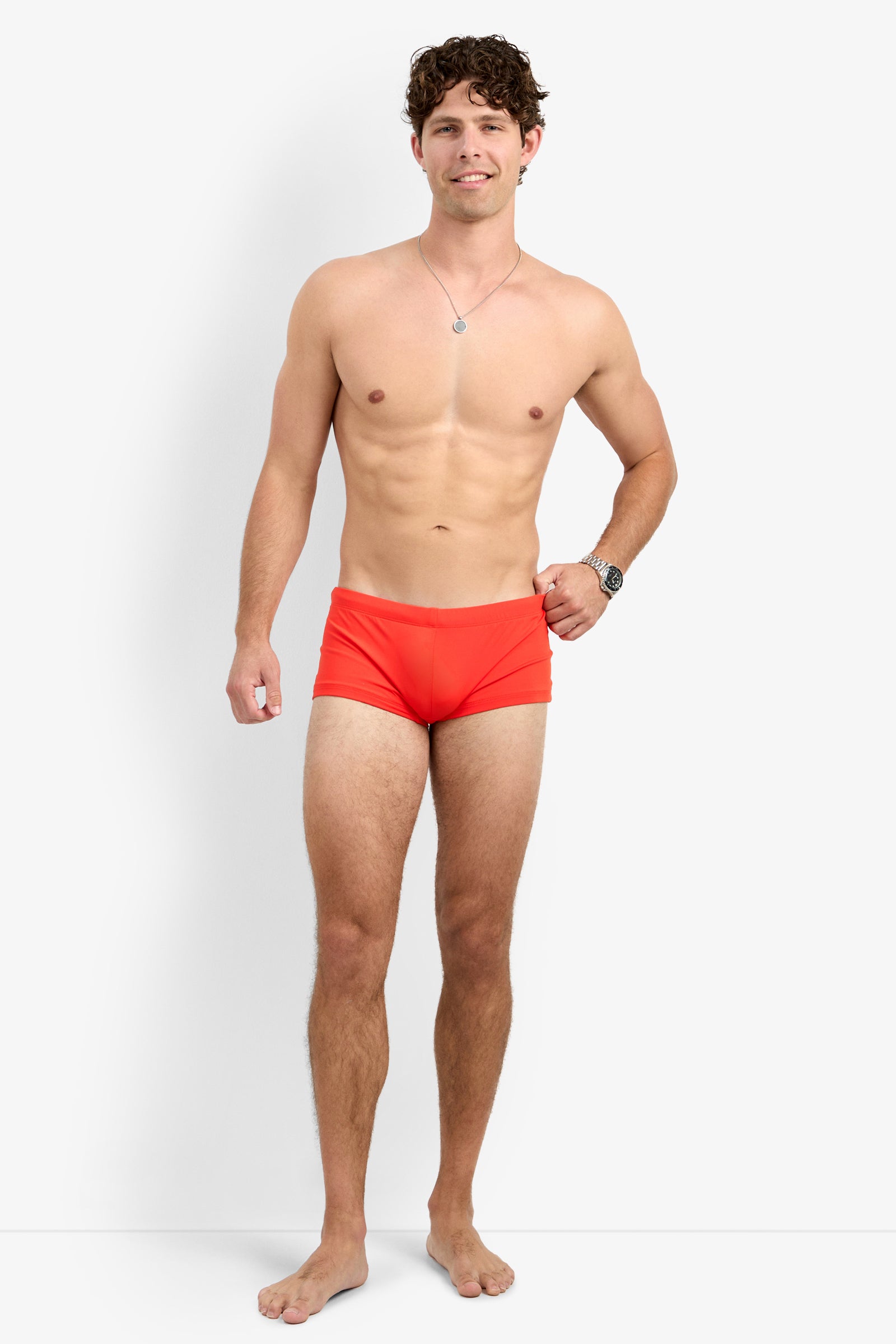 A smiling man stands barefoot against a white background, confidently posing in the Montego Bay Swim Trunk, paired with a wristwatch and necklace. One hand rests relaxed by his side.