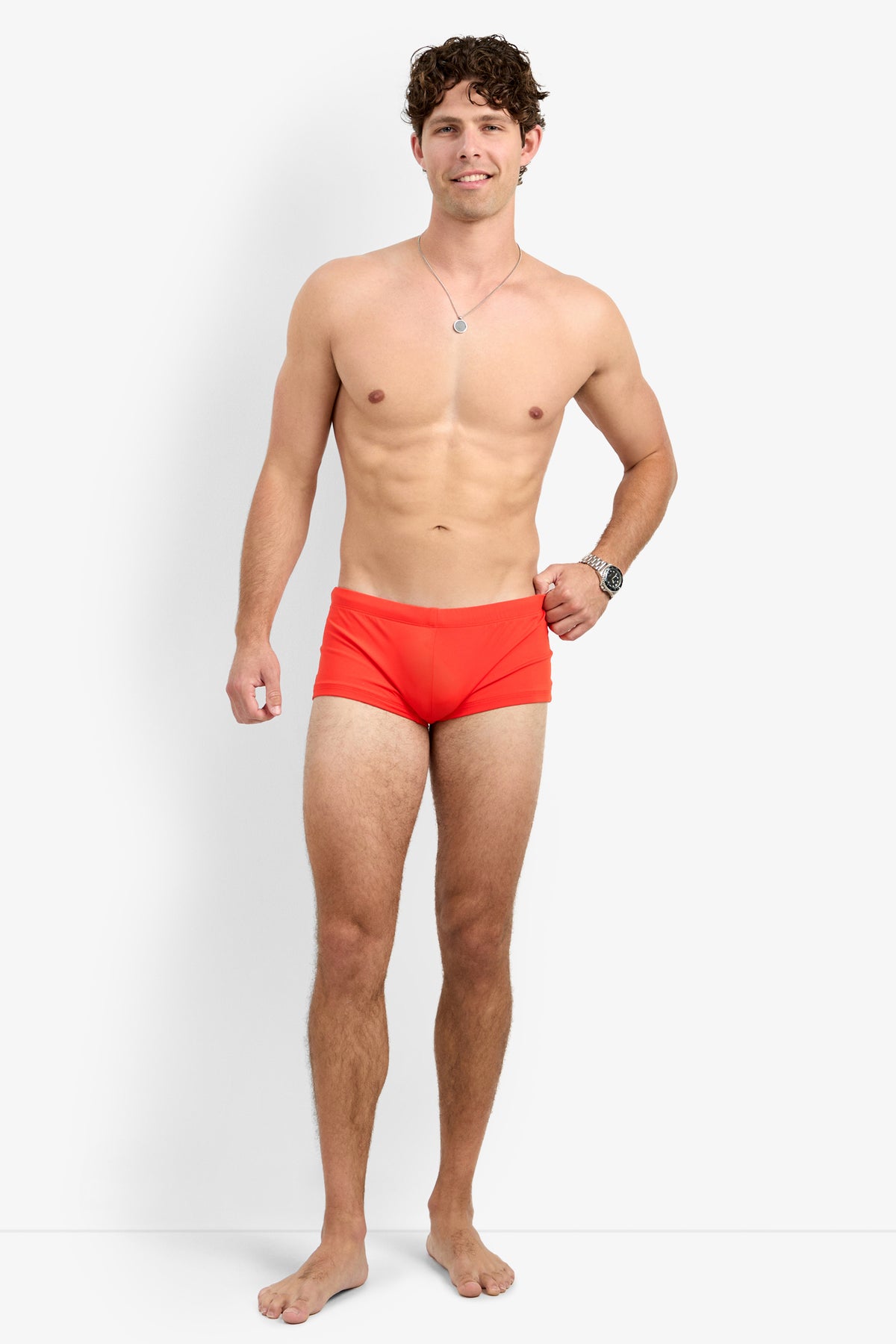 A smiling man stands barefoot against a white background, confidently posing in the Montego Bay Swim Trunk, paired with a wristwatch and necklace. One hand rests relaxed by his side.