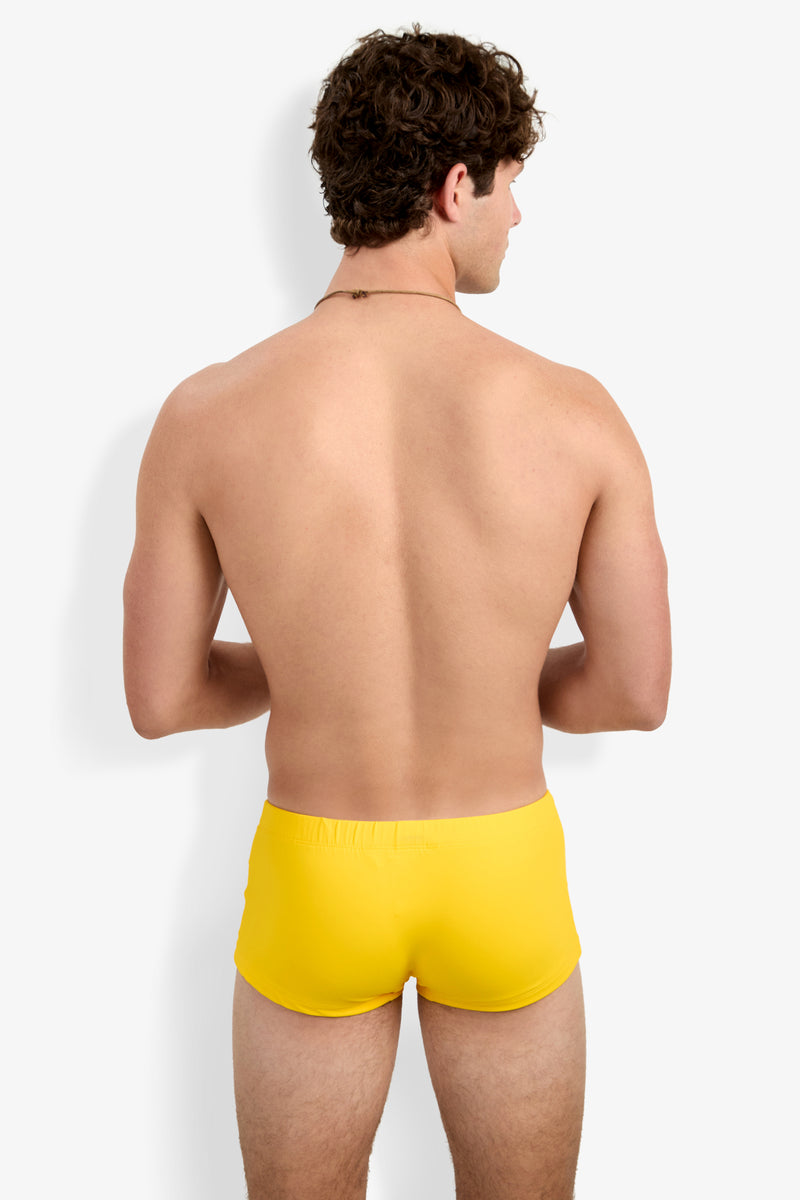 A shirtless man with short curly hair, facing away from the camera, models the Montego Bay Swim Trunk in bright yellow against a plain white background.