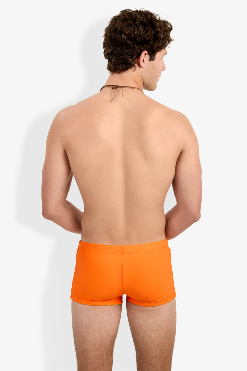 A man with curly brown hair, seen from behind, wears Seaside Beach Swim Trunk in bright orange and a thin cord necklace against a plain white background.