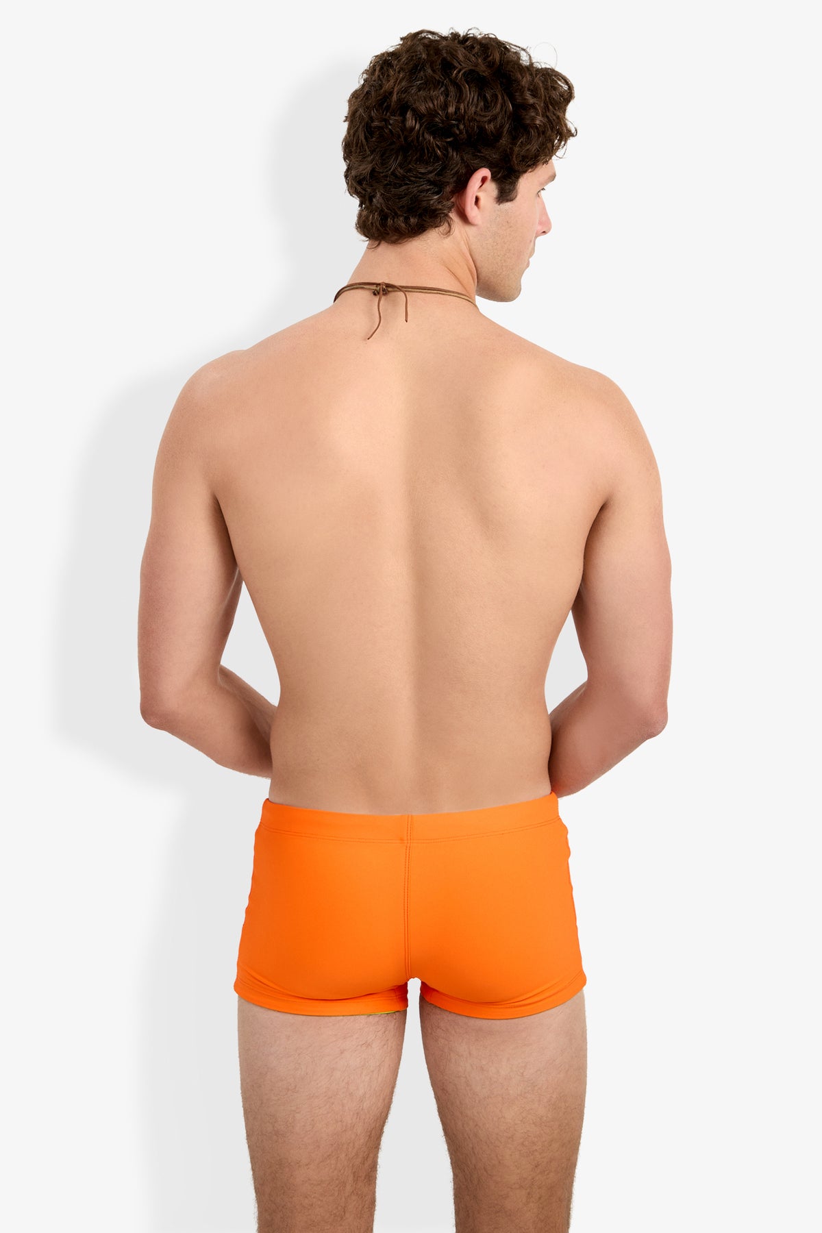 A man with curly brown hair faces away from the camera, modeling Seaside Beach Swim Trunk in bright orange and a string-tie necklace against a plain white background.