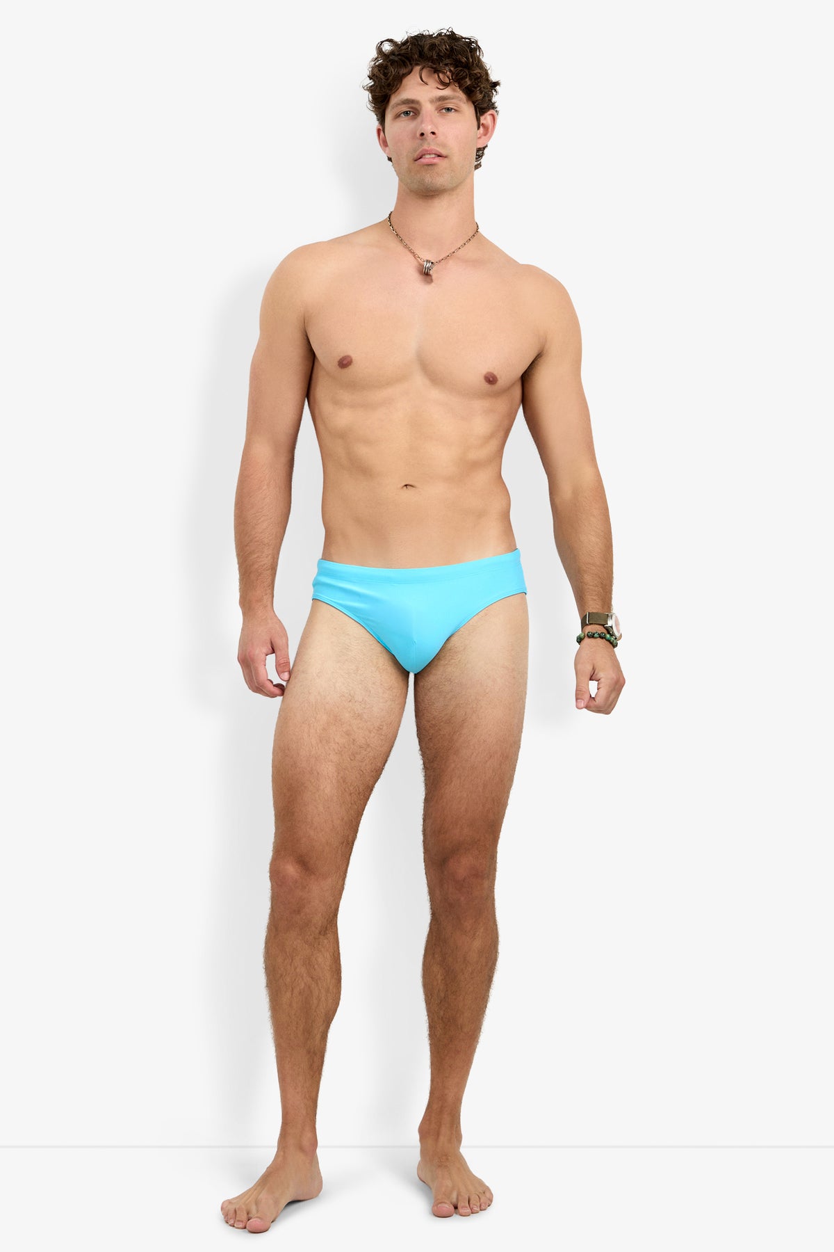 A man with short curly hair and a neutral expression stands barefoot against a white background, wearing only the Palm Beach Swim Brief, a necklace, and a wristwatch.