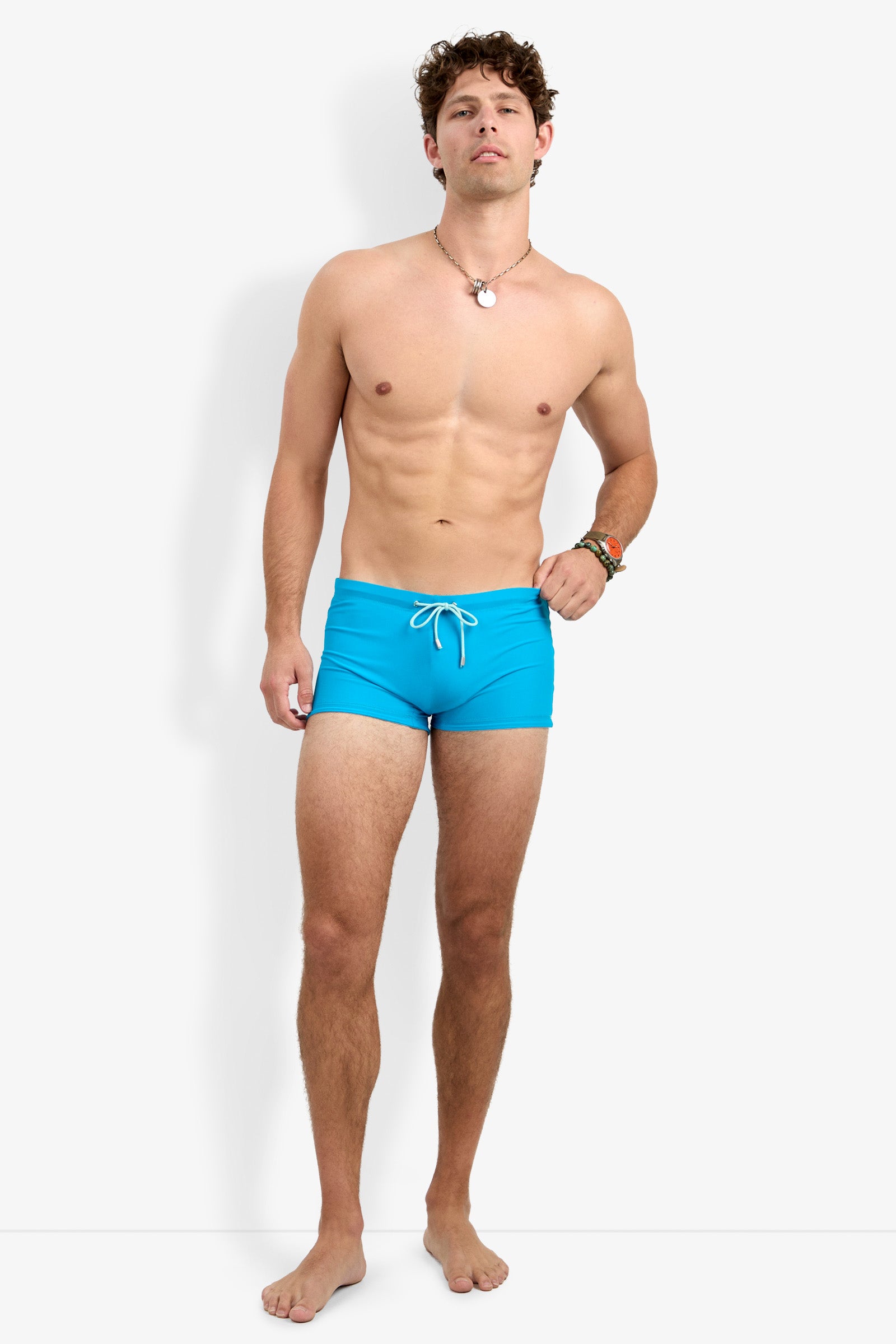 A young man stands barefoot against a white background, wearing the Seaside Beach Swim Trunk in bright blue with a white drawstring. Shirtless and accessorized with a necklace and bracelet, he gazes slightly upward with a neutral expression.