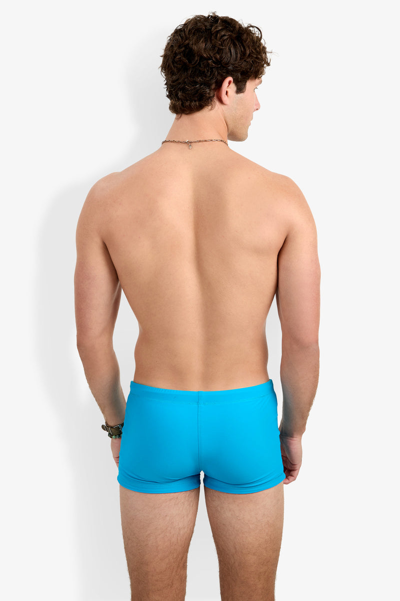 A man with curly brown hair is seen from behind, shirtless, wearing Seaside Beach Swim Trunk in bright blue, set against a plain white background.