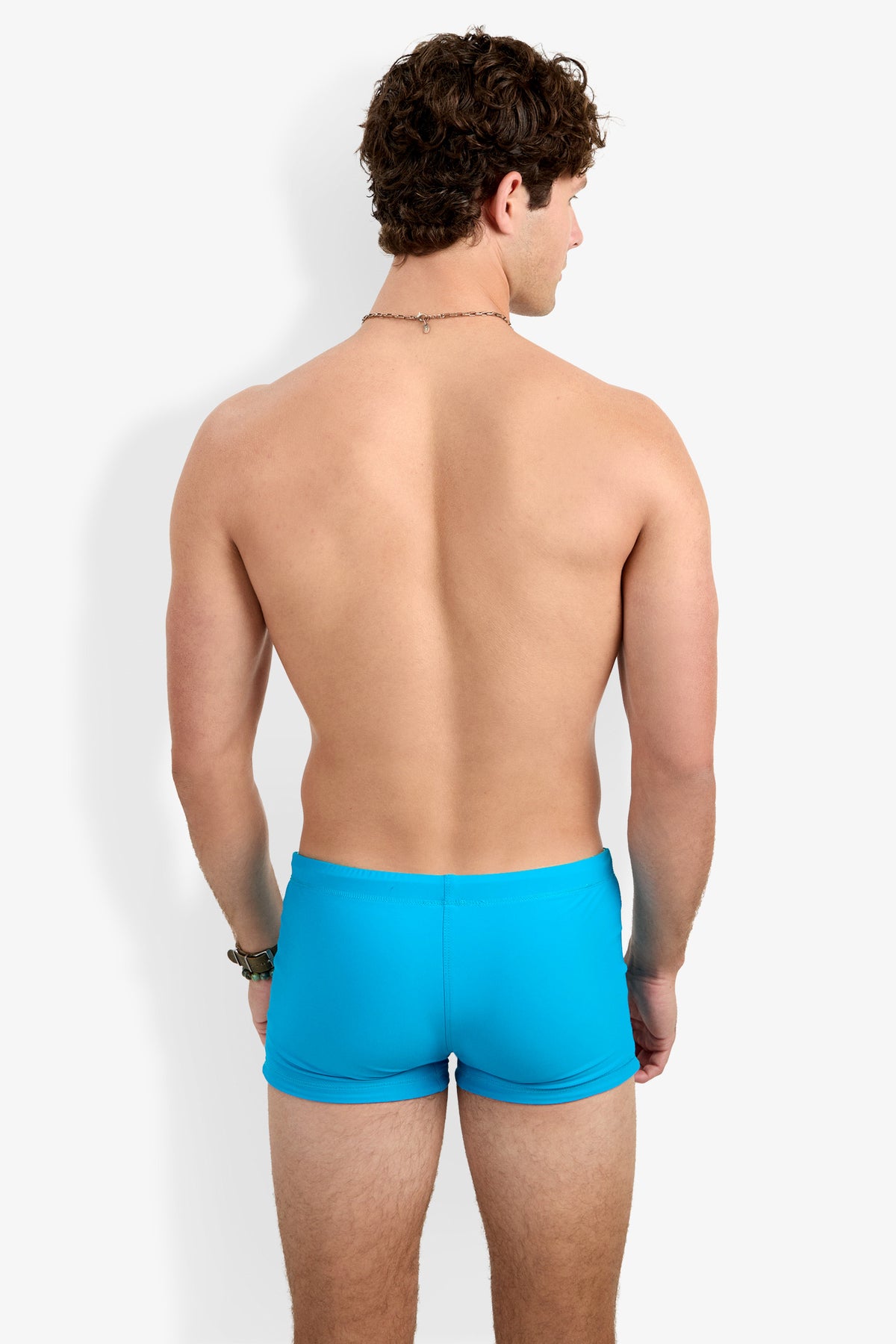 A man with curly brown hair stands with his back to the camera, wearing Seaside Beach Swim Trunk in bright blue. He is shirtless and faces a plain white background.
